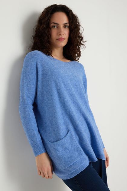 A person with curly brown hair wears the Two Pocket Oversize Knit—a light blue, oversized women's knitwear jumper with front pockets—and dark pants, standing against a white wall.