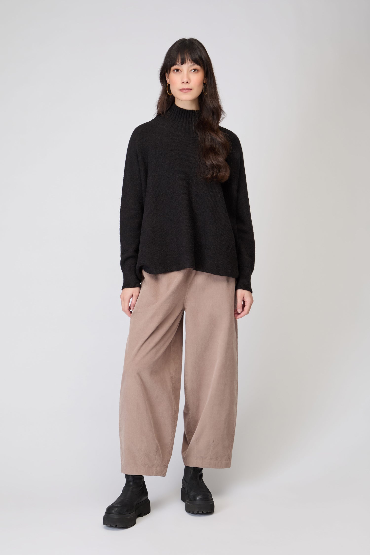 A person stands facing forward, wearing the Boxy High Neck Knit in black, wide-leg beige pants, and black boots against a plain light background.