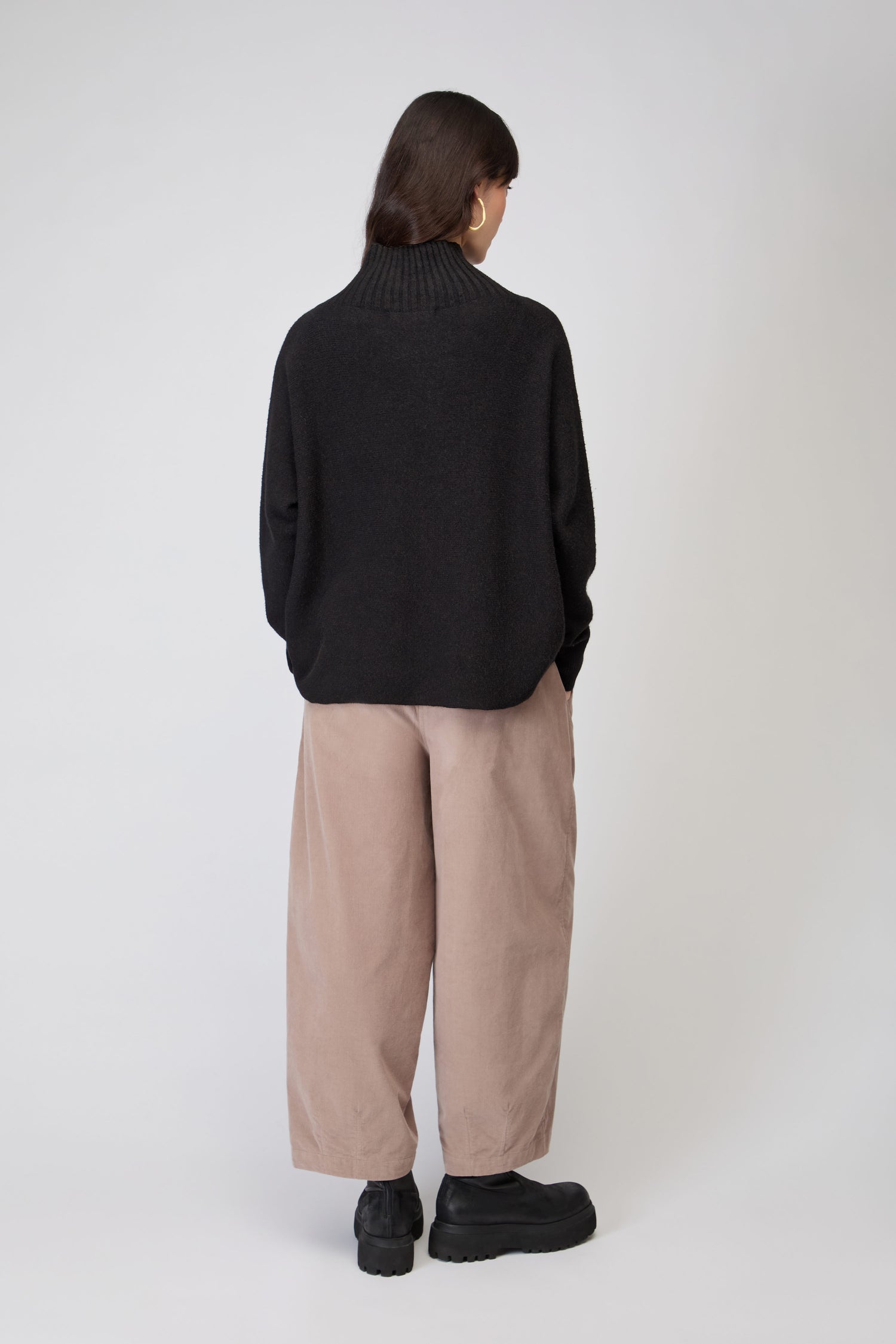 A person stands facing away from the camera, wearing the Boxy High Neck Knit in black, beige wide-leg pants, and black shoes against a plain light background.