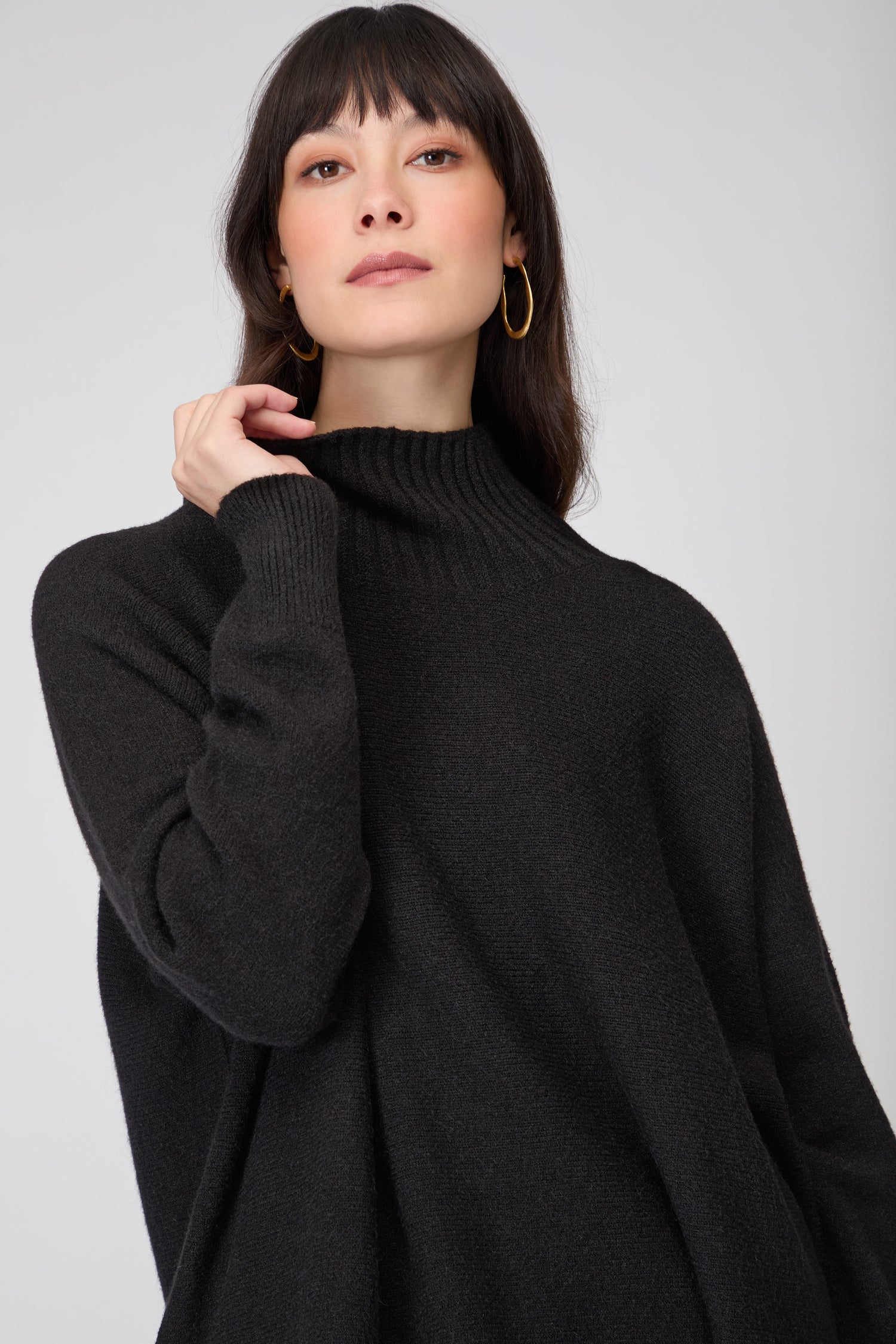 A woman with long dark hair wears the Boxy High Neck Knit in black and gold hoop earrings, posing against a plain light background.