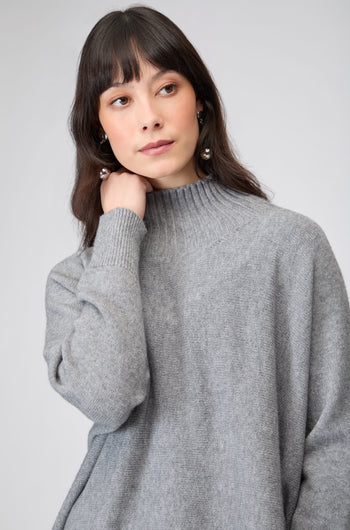 A woman with long brown hair wears the Boxy High Neck Knit in gray, looking slightly to the side with a neutral expression.