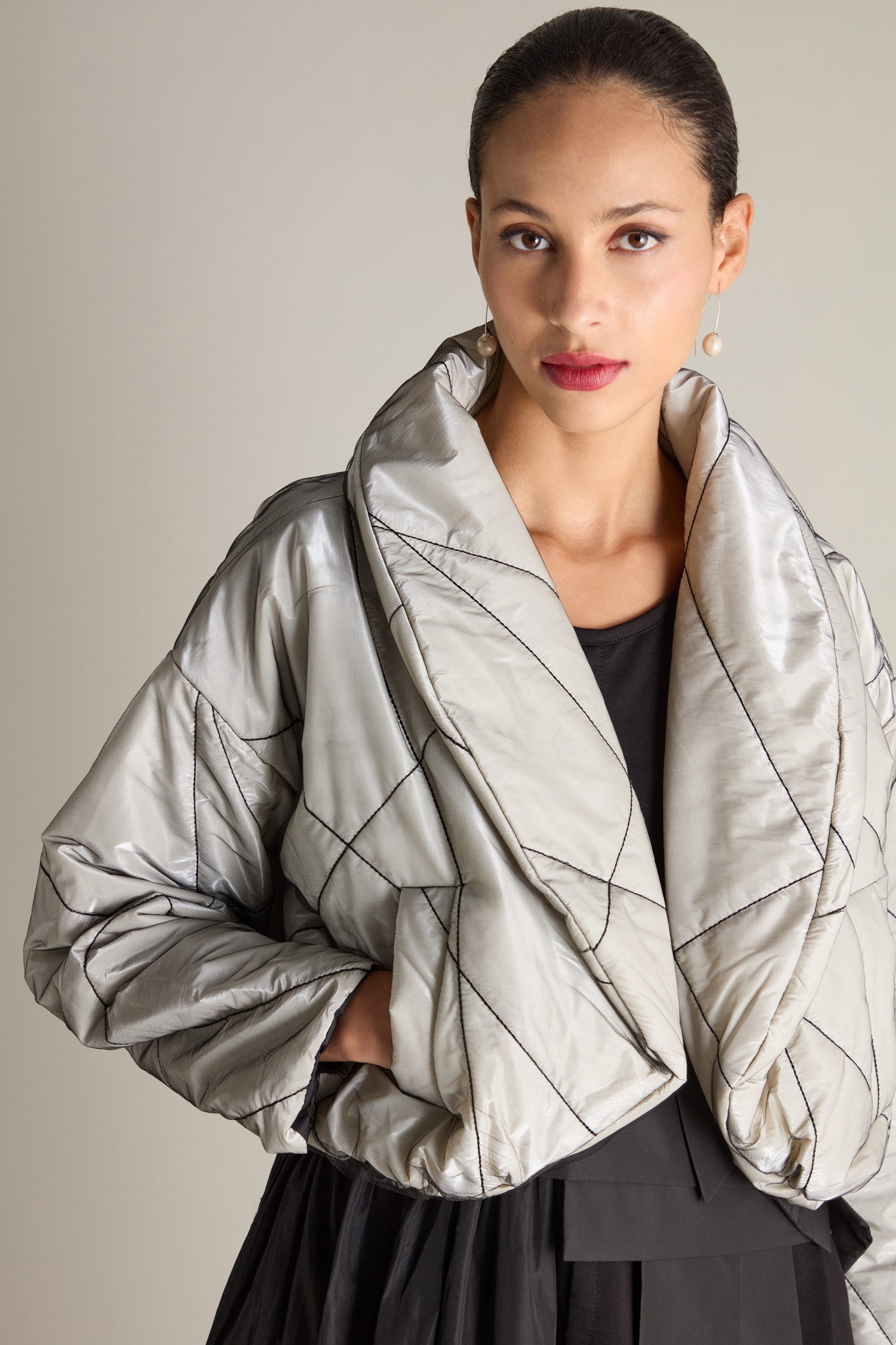 A woman stands against a plain background, adorned in a Short Puffa Evening Jacket. The silver, quilted short puffa jacket features a high collar. With her hands tucked into the pockets and her gaze directed at the camera, the jacket's curved hem adds a subtle touch of style to her ensemble.