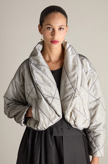 A woman with pulled-back hair is wearing the Short Puffa Evening Jacket, a quilted, metallic silver jacket featuring a large collar and black stitching, over a black outfit. She stands against a plain backdrop, gazing forward.