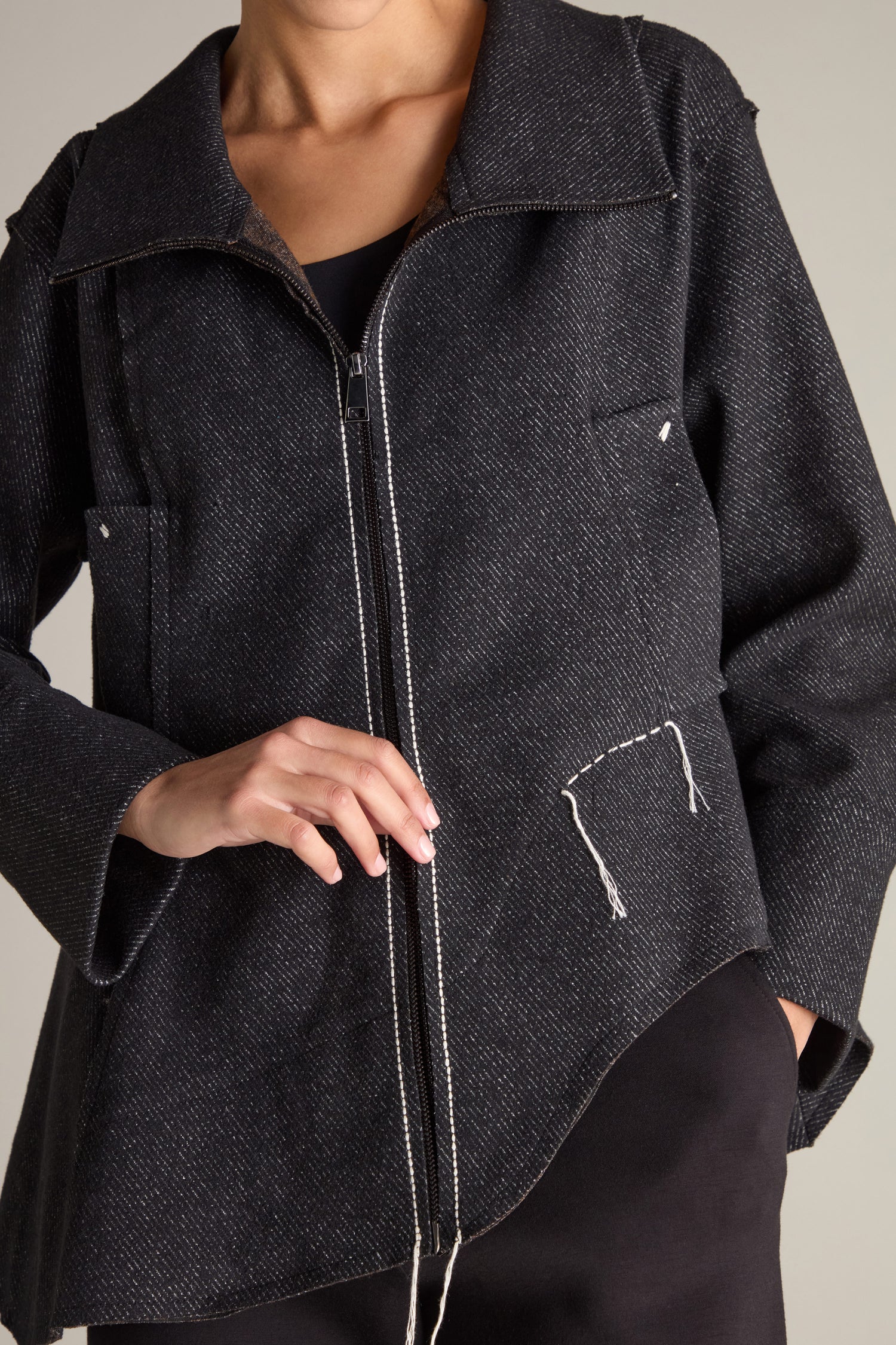 A person wearing the Zip Detail Jacket, which features visible stitches and a zipper, is holding the fabric with one hand. The jacket boasts oversized front pockets and an unfinished, rough design with deconstructed edges. The background is plain and neutral.