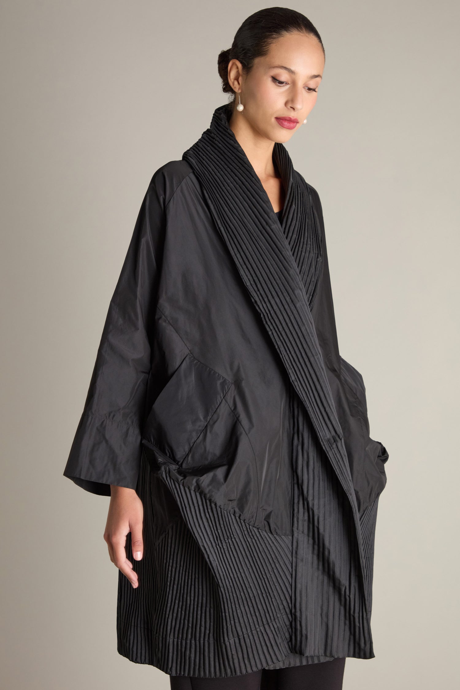 A woman is standing against a plain background wearing a loose, black Rib Detail Evening Coat with pleated details and two large front pockets. The kimono-style front adds elegance to her look. She has her hair pulled back and is looking down.