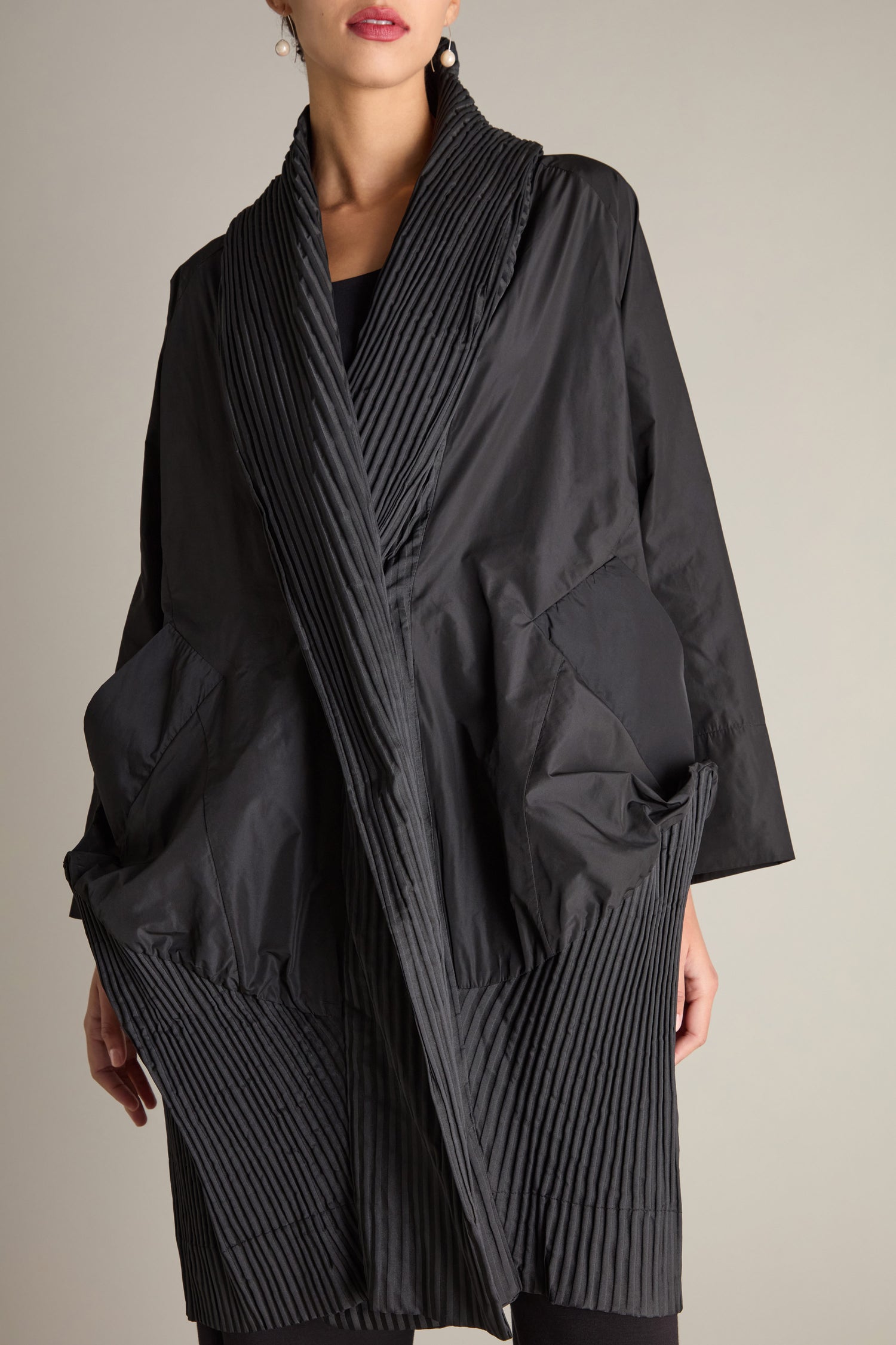 A person wearing the Rib Detail Evening Coat, characterized by its black color and loose-fitting design, stands against a plain beige background. The long-line silhouette complements the kimono-style front, with their face partially out of the frame.