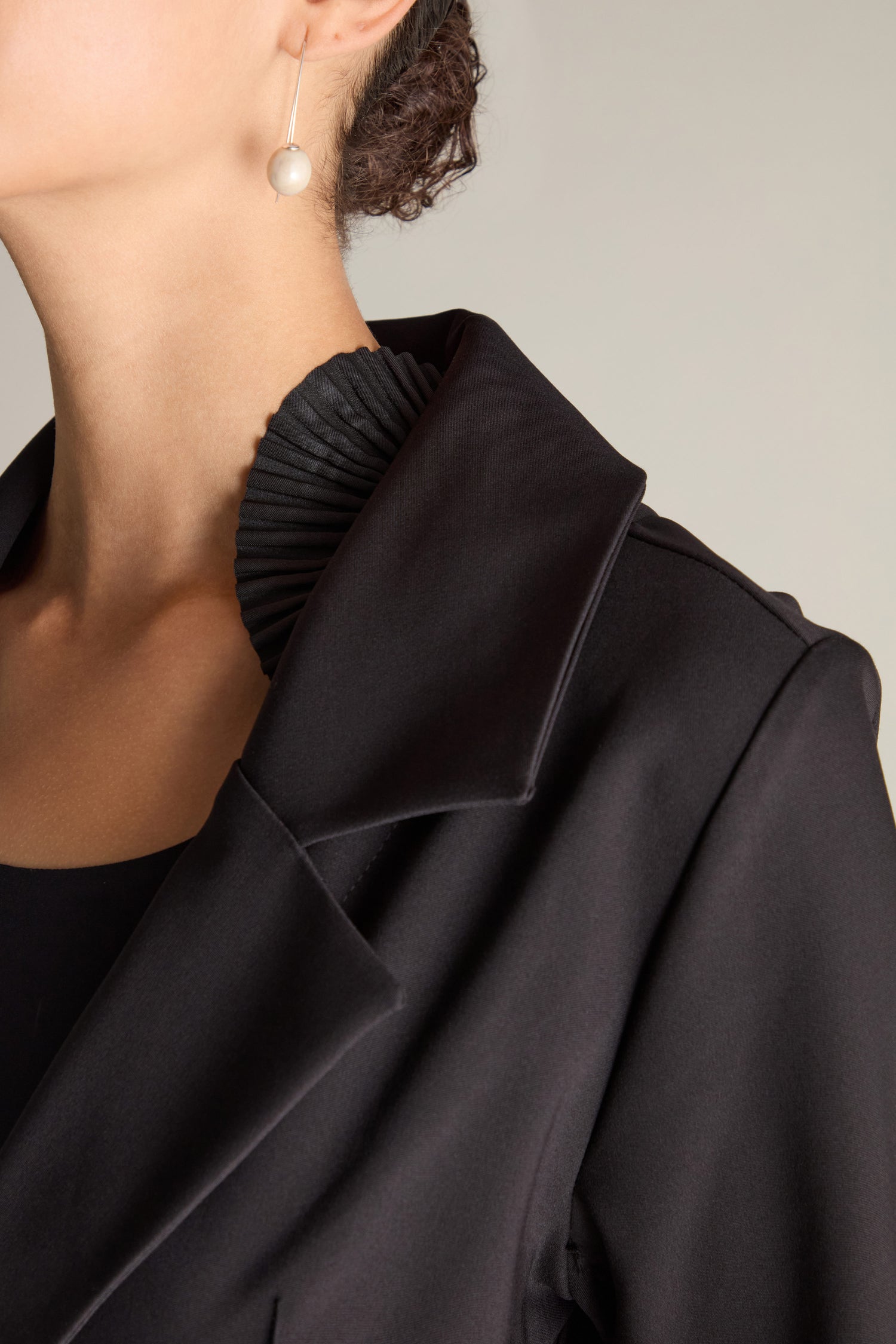 Close-up of a woman’s upper body in a black Fitted Fan Detail Jacket, featuring a classic lapel and 3D fan frills applique. Her face is partially visible and adorned with a pearl earring.