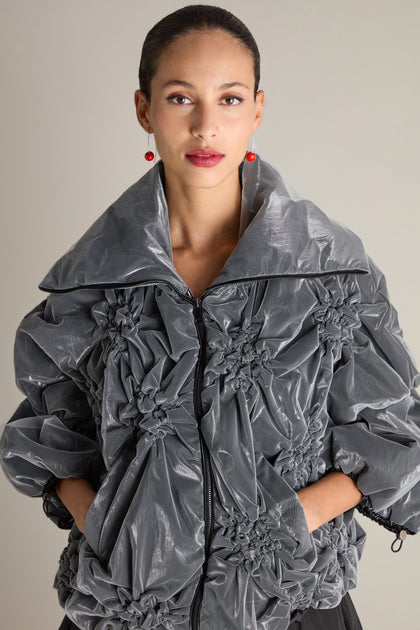 A woman with pulled-back hair stands against a neutral background. She wears the Ruched Collar Zip Jacket in grey, featuring ornate fabric detailing and pairs it with red spherical earrings. With her hands casually in her pockets, she makes it clear that her outfit is a contemporary statement piece.