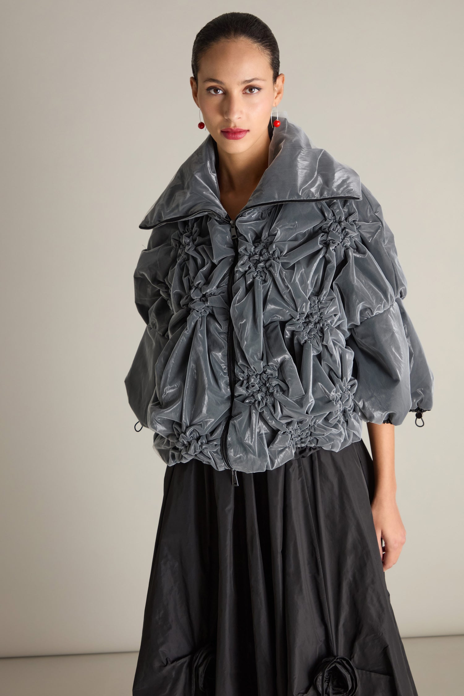 A person stands against a plain background wearing the Ruched Collar Zip Jacket, featuring a gray, textured design with drawstring cuffs, paired with a black skirt, creating a contemporary statement piece.