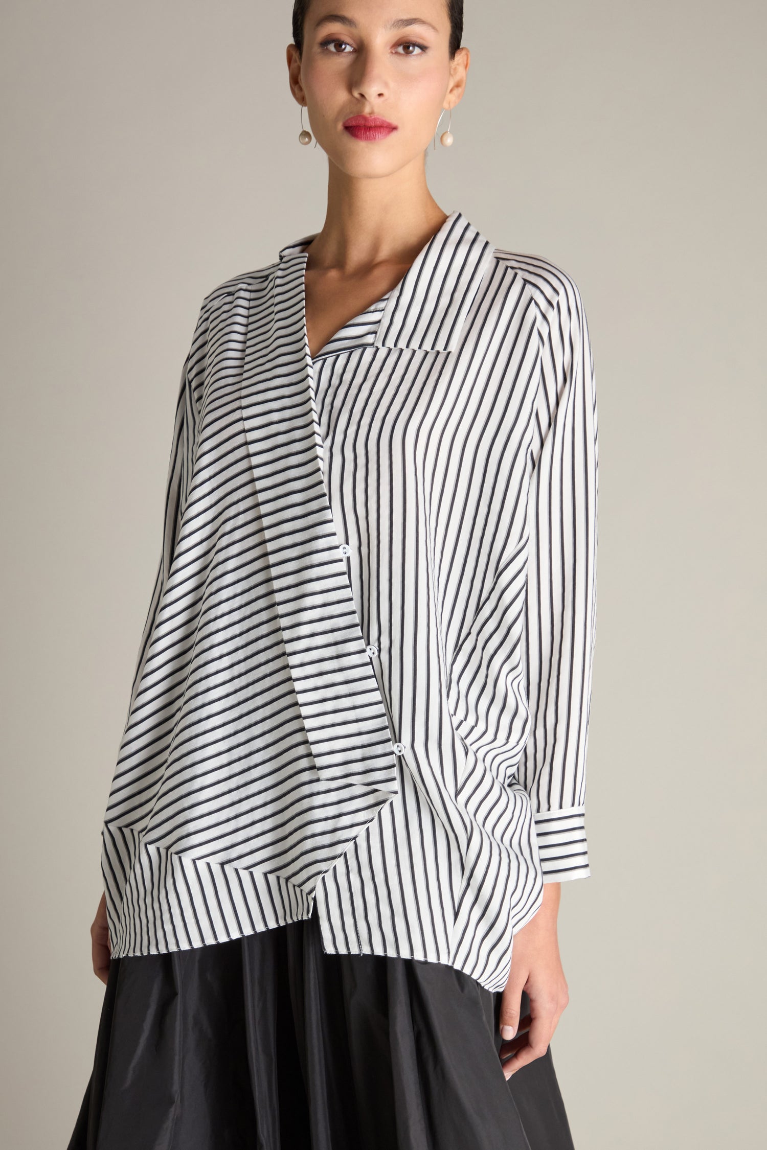 A person wearing the Stripe Asymmetric Shirt, a stylish contemporary black-and-grey striped blouse with asymmetrical draping, paired with a black skirt, stands against a plain background.