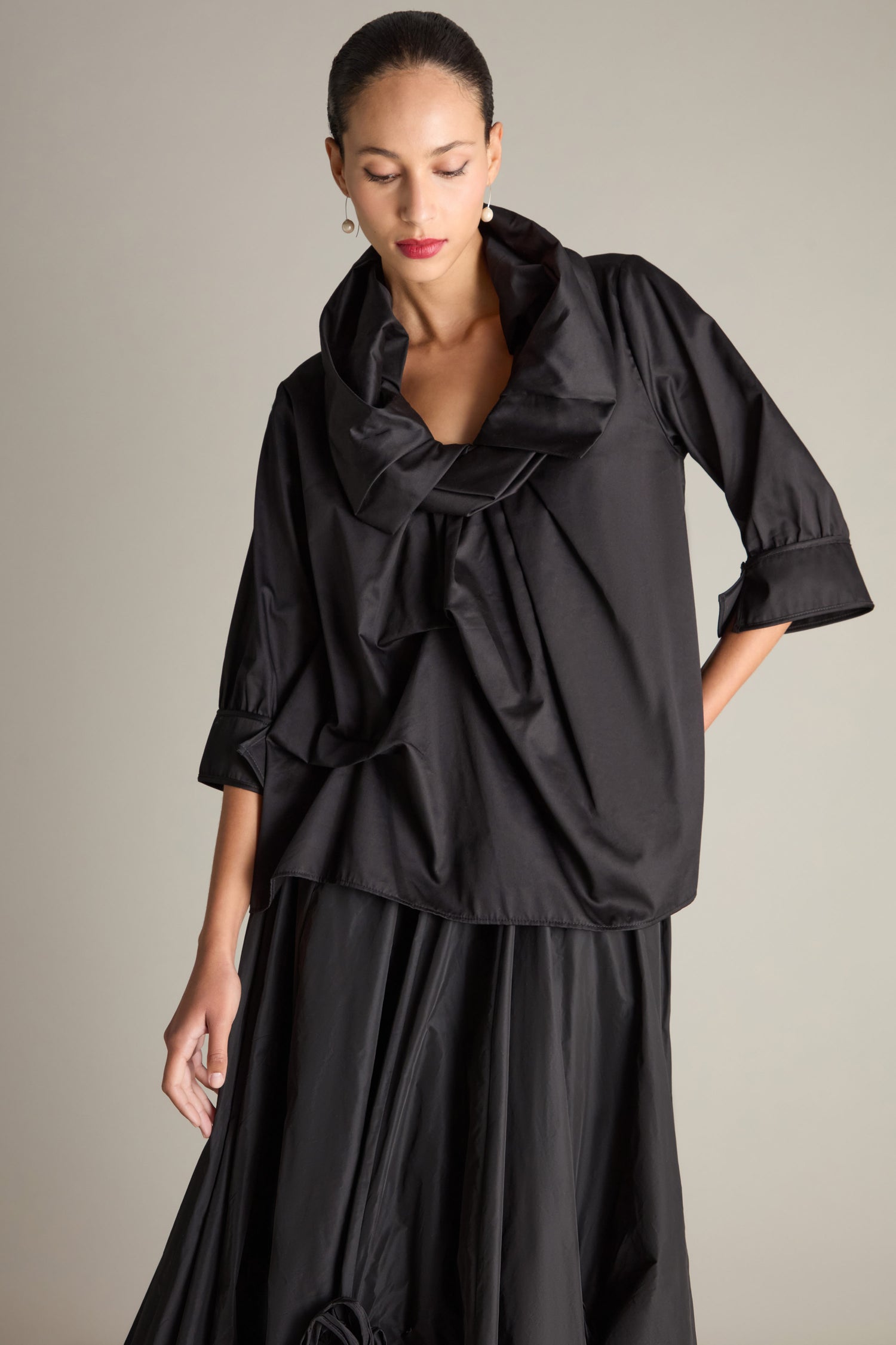 A person wearing the Frill Neck Easy Top, a black blouse with 3/4 sleeves with cuffs and an oversized collar, paired with a black skirt, stands against a neutral background.