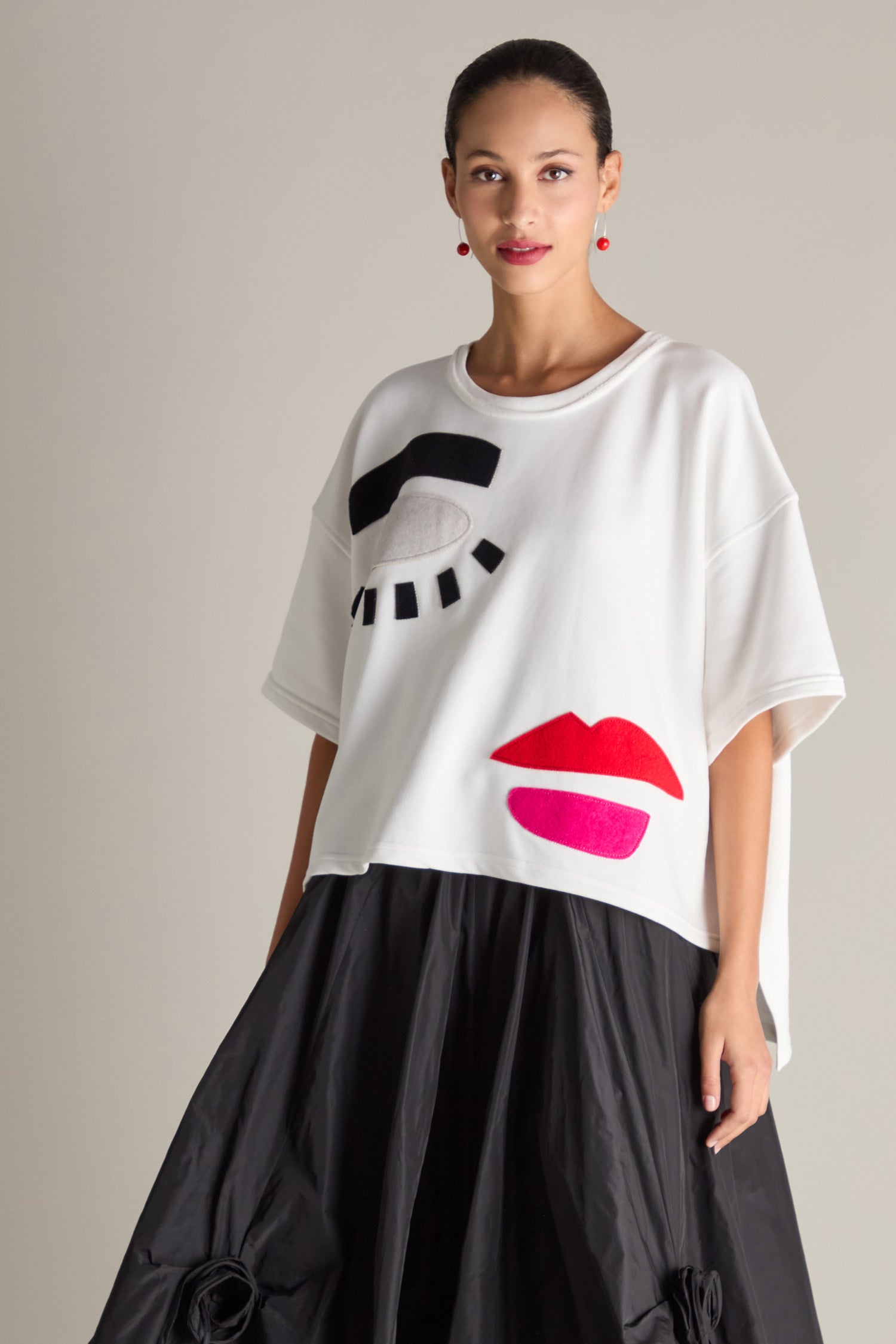 A person is wearing the Eyes And Lips Jersey Top, a white oversized T-shirt embellished with abstract black and red eye and lip designs, paired with a black skirt.