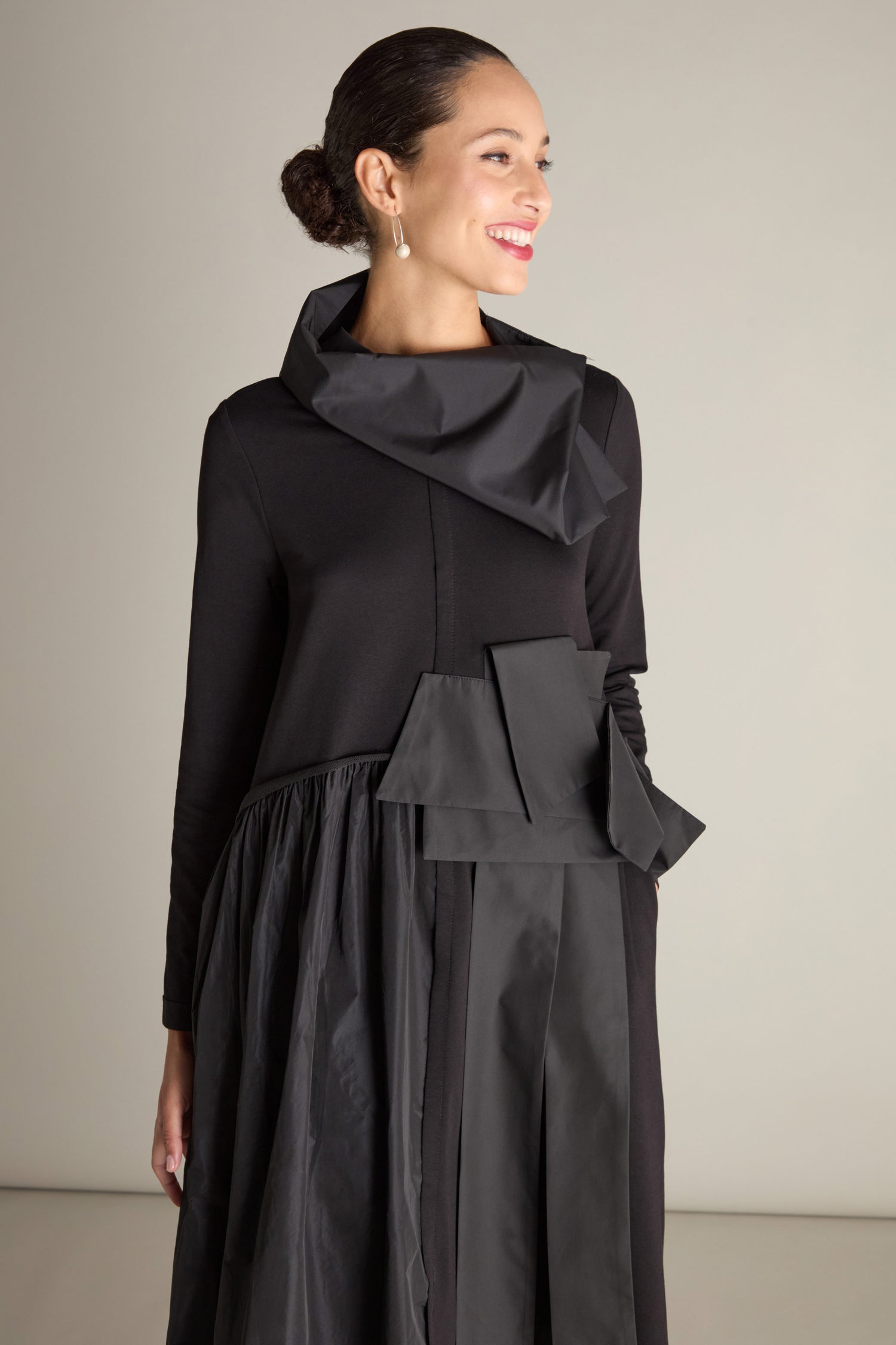 A person wearing the Jersey Taffeta Waist Detail Dress with asymmetrical detailing and 3D taffeta appliqué, standing and smiling, with hair tied back.