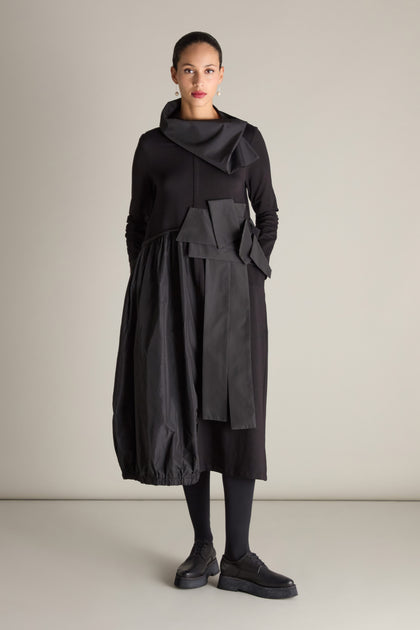 A person stands against a plain background wearing a Jersey Taffeta Waist Detail Dress in black, featuring an asymmetrical design with large pockets, paired with matching black shoes.