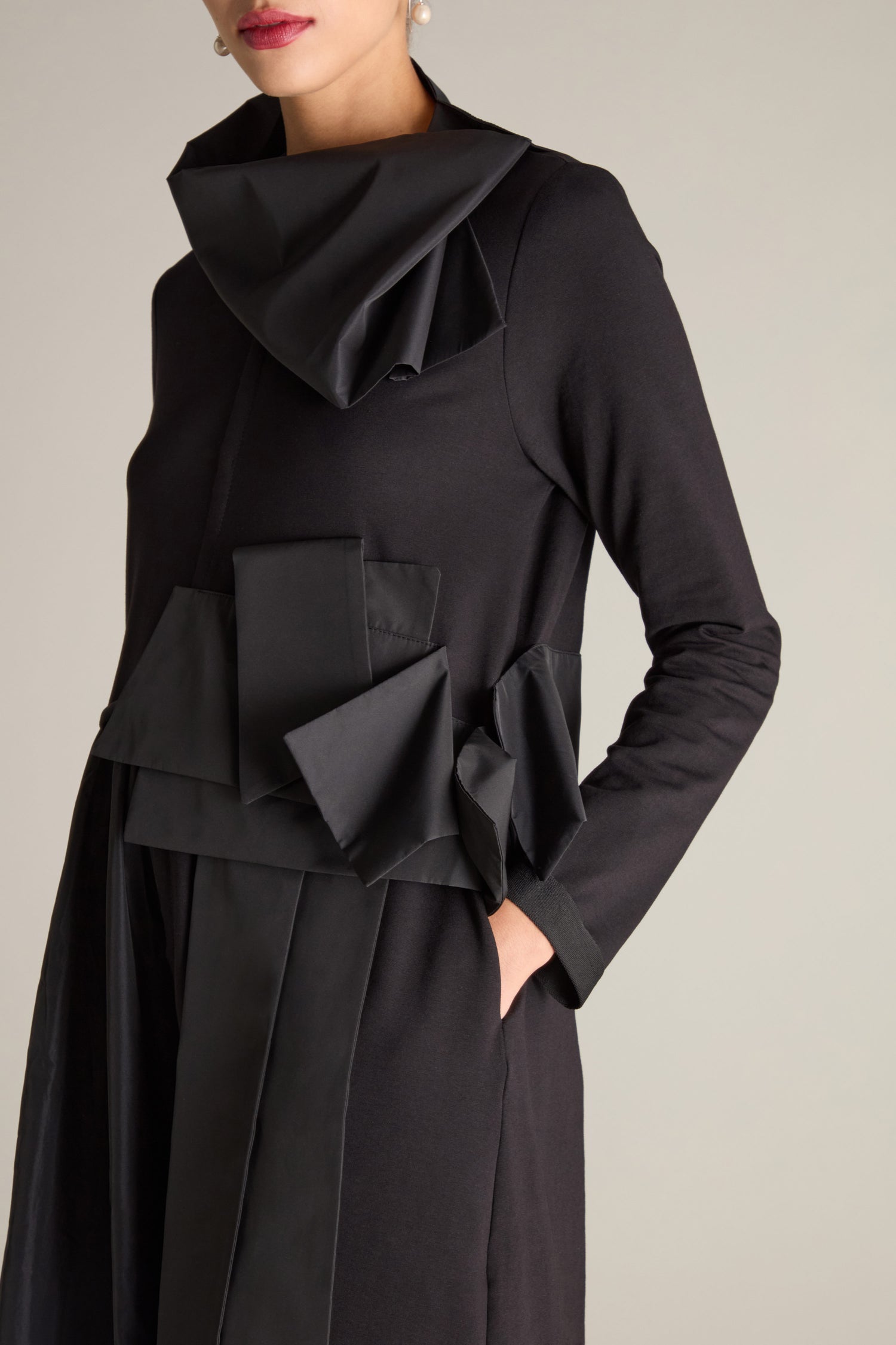 A person wearing the Jersey Taffeta Waist Detail Dress, which features long sleeves, a black color, and an asymmetrical collar. The dress includes decorative fabric folds around the waist and pockets, highlighted by a subtle 3D taffeta appliqué that adds texture.