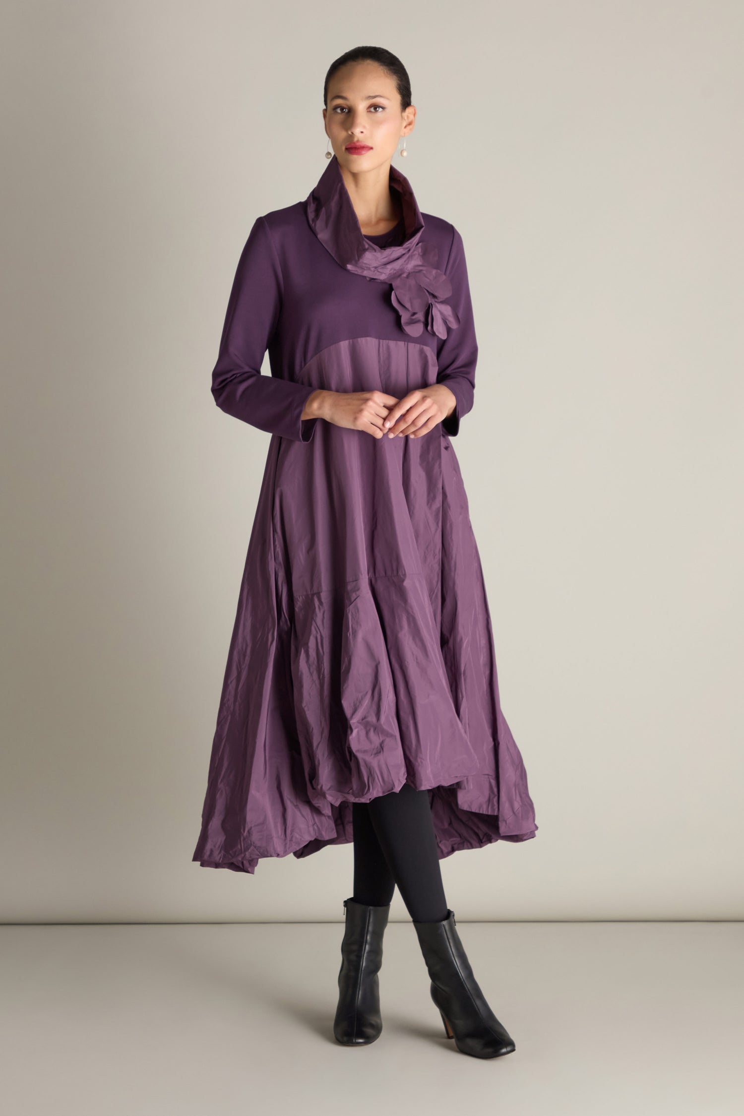 A person is dressed in a purple Jersey Taffeta Flower Dress featuring long sleeves, an asymmetrical design, and an empire waistline. They have paired it with black leggings and ankle boots while posing against a plain background.