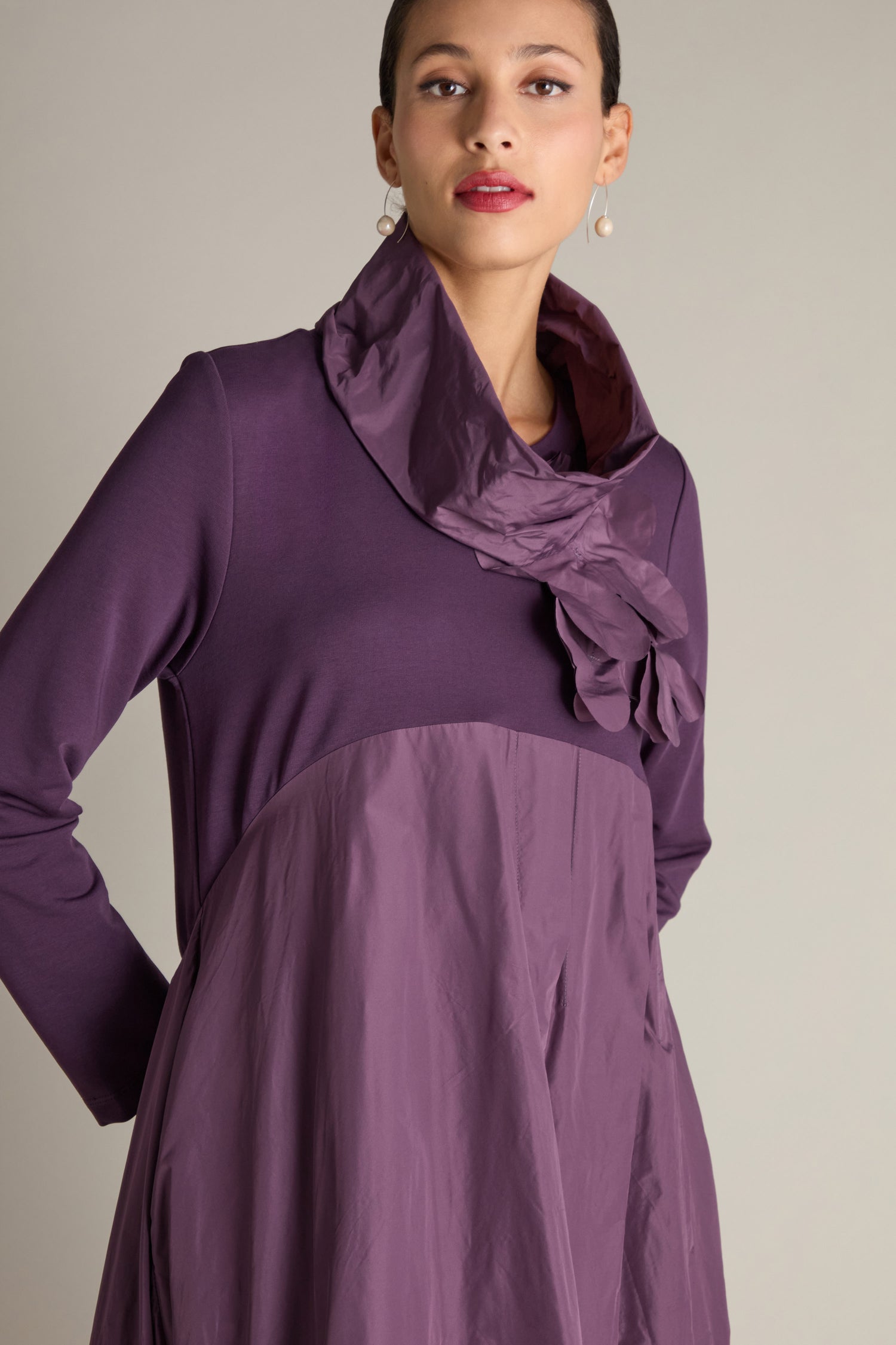 A person stands against a neutral background, wearing the Jersey Taffeta Flower Dress in purple, which showcases a high collar and delicate flower details. This elegant ensemble is completed with an A-line puffball skirt that enhances its charm.