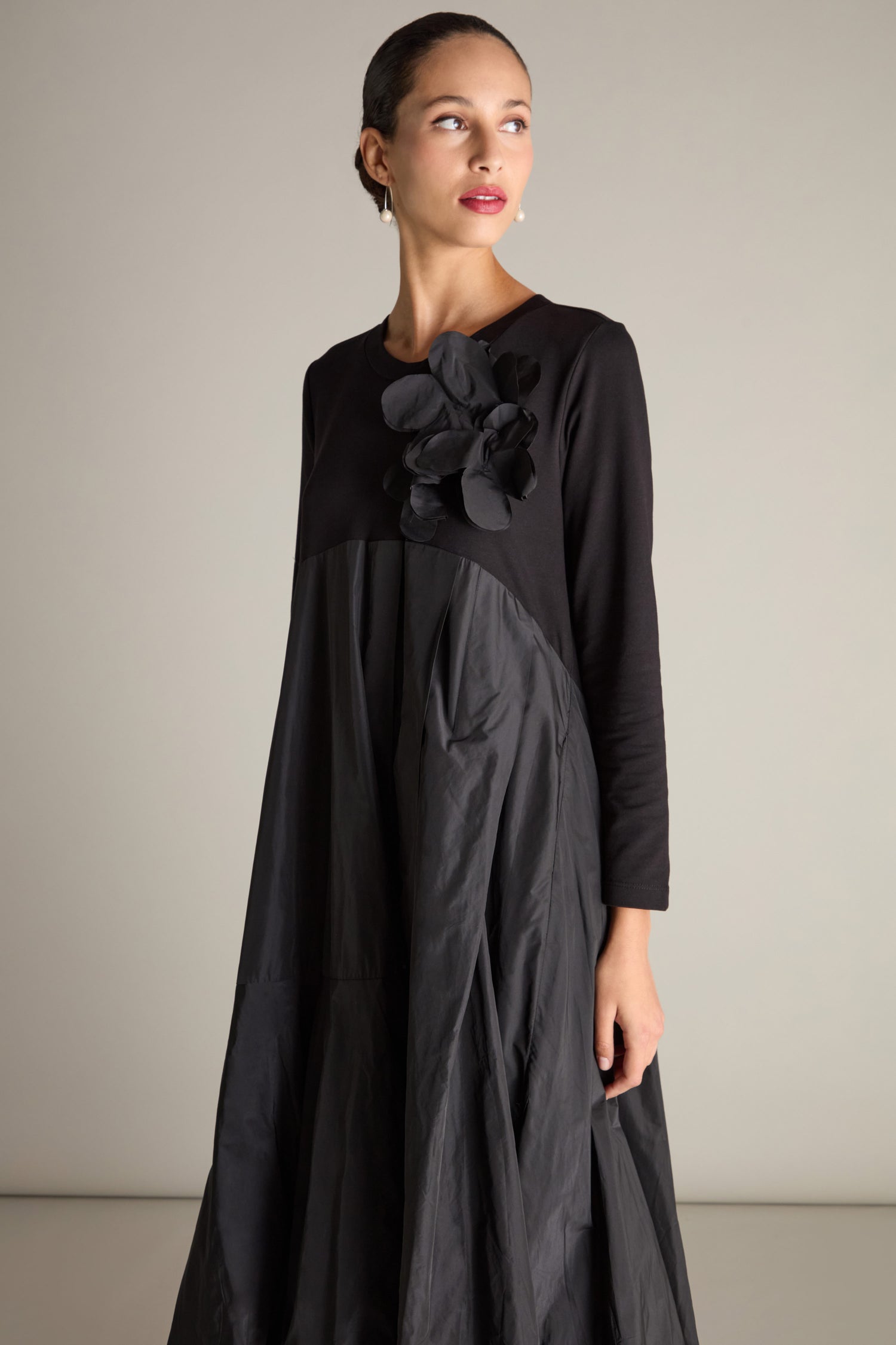 In a neutral setting, an individual is dressed in the Jersey Taffeta Flower Dress, a long-sleeve black outfit adorned with floral embellishments on the chest and an empire waistline, while gazing to the side.