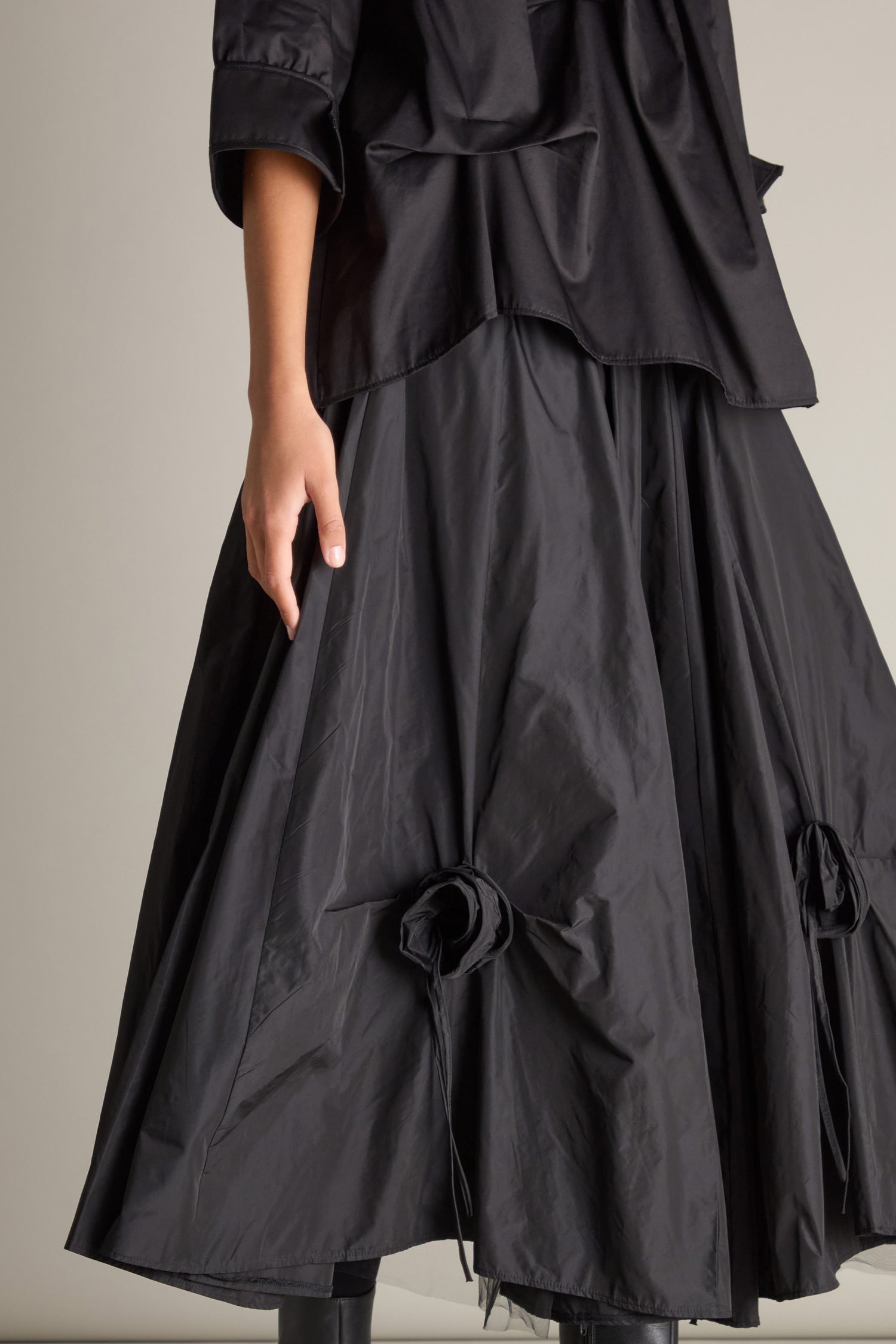 A person attired in a sophisticated black ensemble, showcasing the Taffeta Flower Detail Skirt adorned with gathered, 3D taffeta flower appliques near the hem.