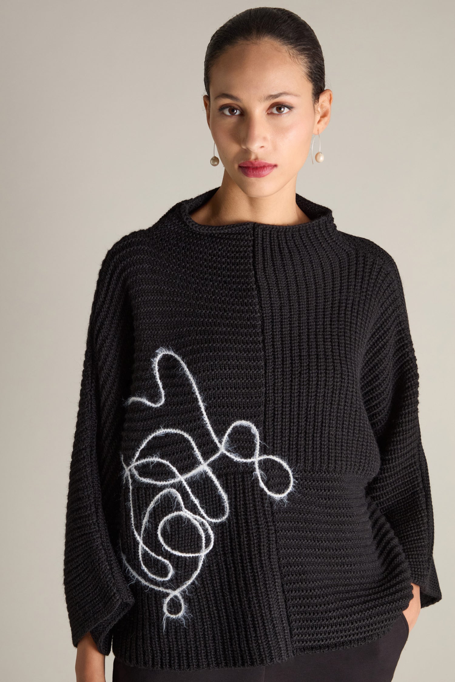 A woman is adorned in the Topstitch Detail Rib Knit, a black knitted sweater showcasing a white abstract design on the front. The piece is crafted from rib knit fabric and includes topstitch details. She complements her outfit with gold earrings while standing against a plain background.