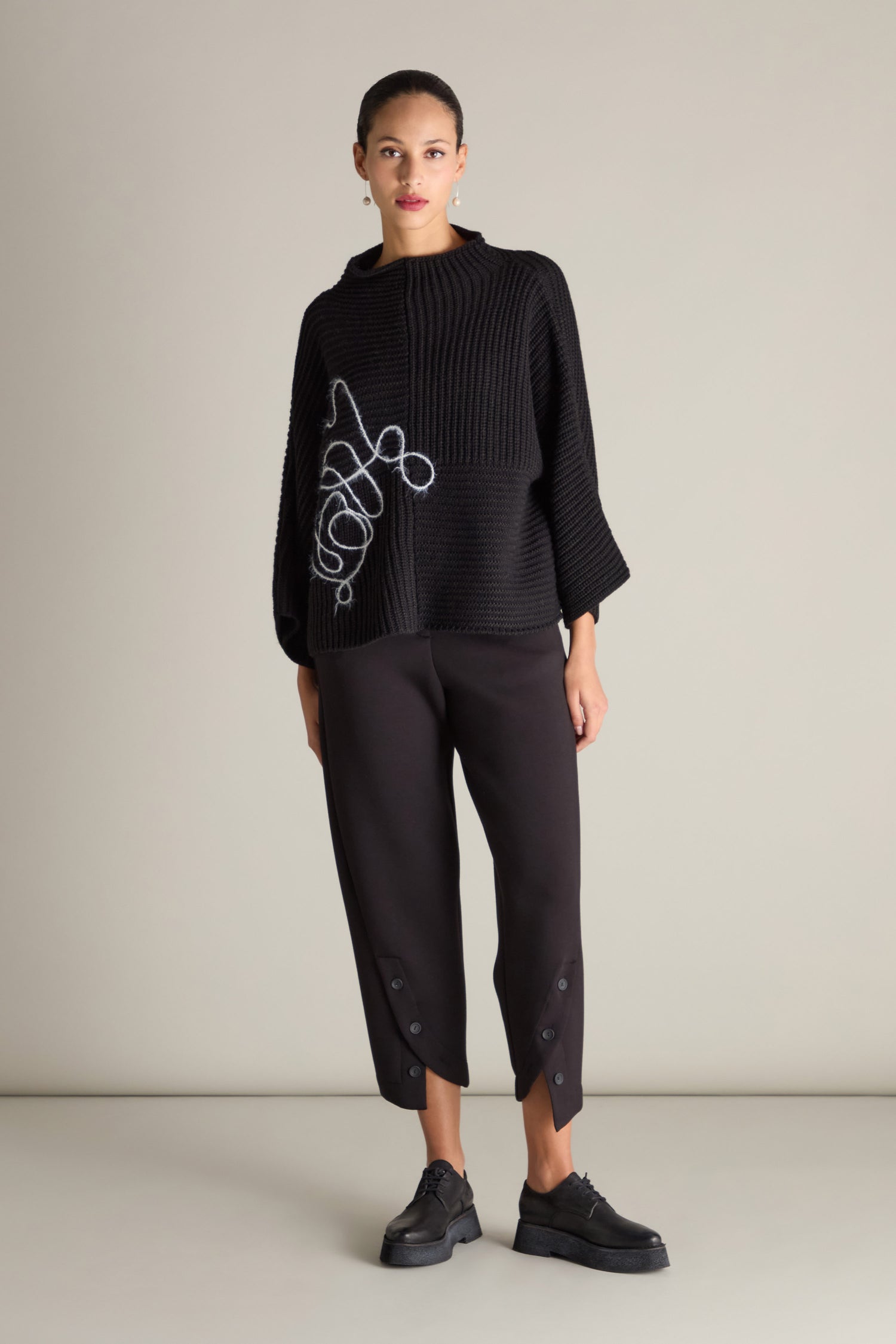 A person stands against a plain background, wearing the Topstitch Detail Rib Knit—a black textured sweater with white abstract embroidery and matching black pants featuring topstitch detail near the hem. The ensemble is completed with black shoes, enhancing the relaxed silhouette provided by the sweater’s rib knit fabric.