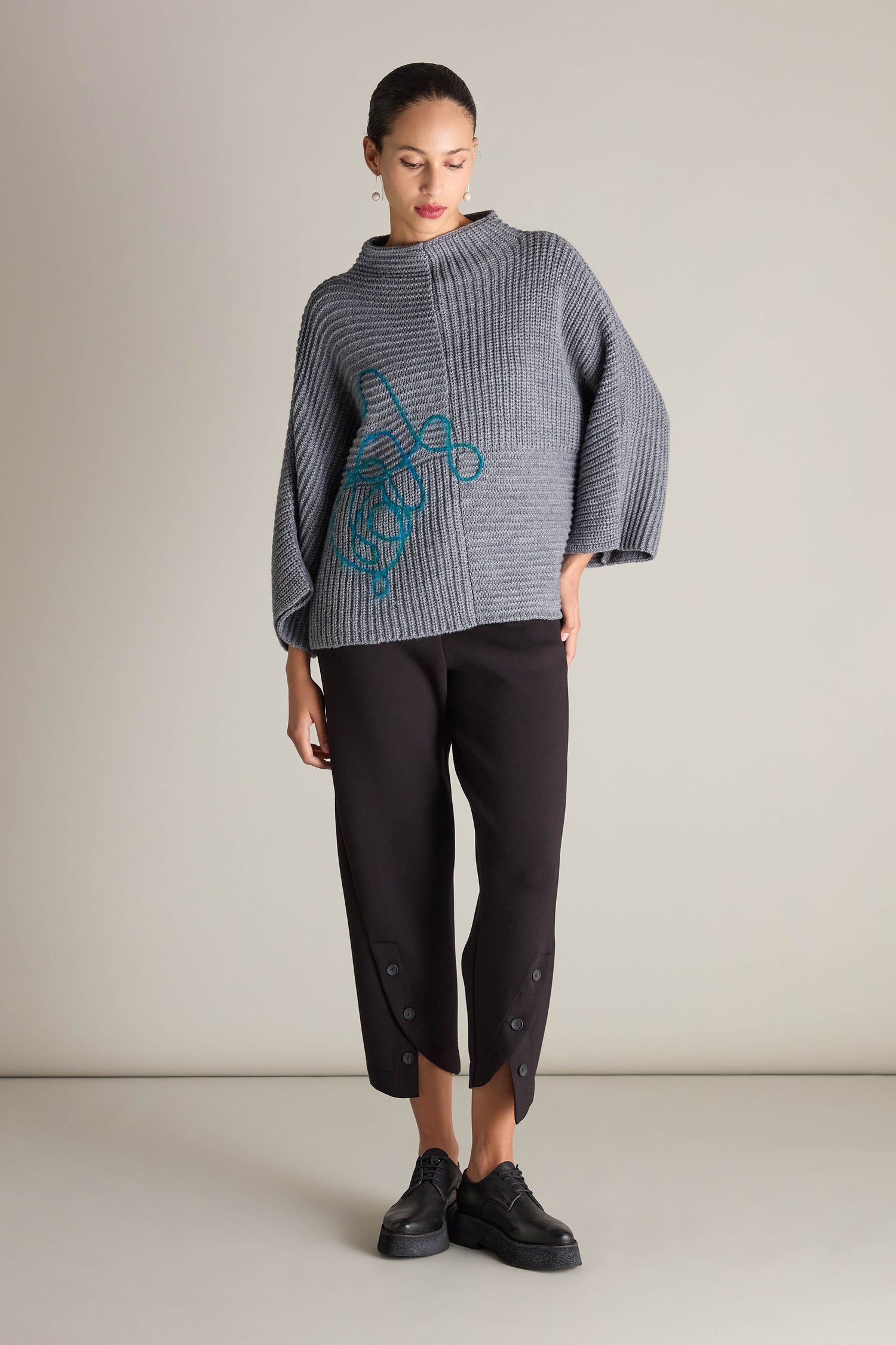 Standing against a plain background, the person is wearing the Topstitch Detail Rib Knit, a textured gray sweater with a blue design crafted from premium fabric, paired with black buttoned pants and black shoes.