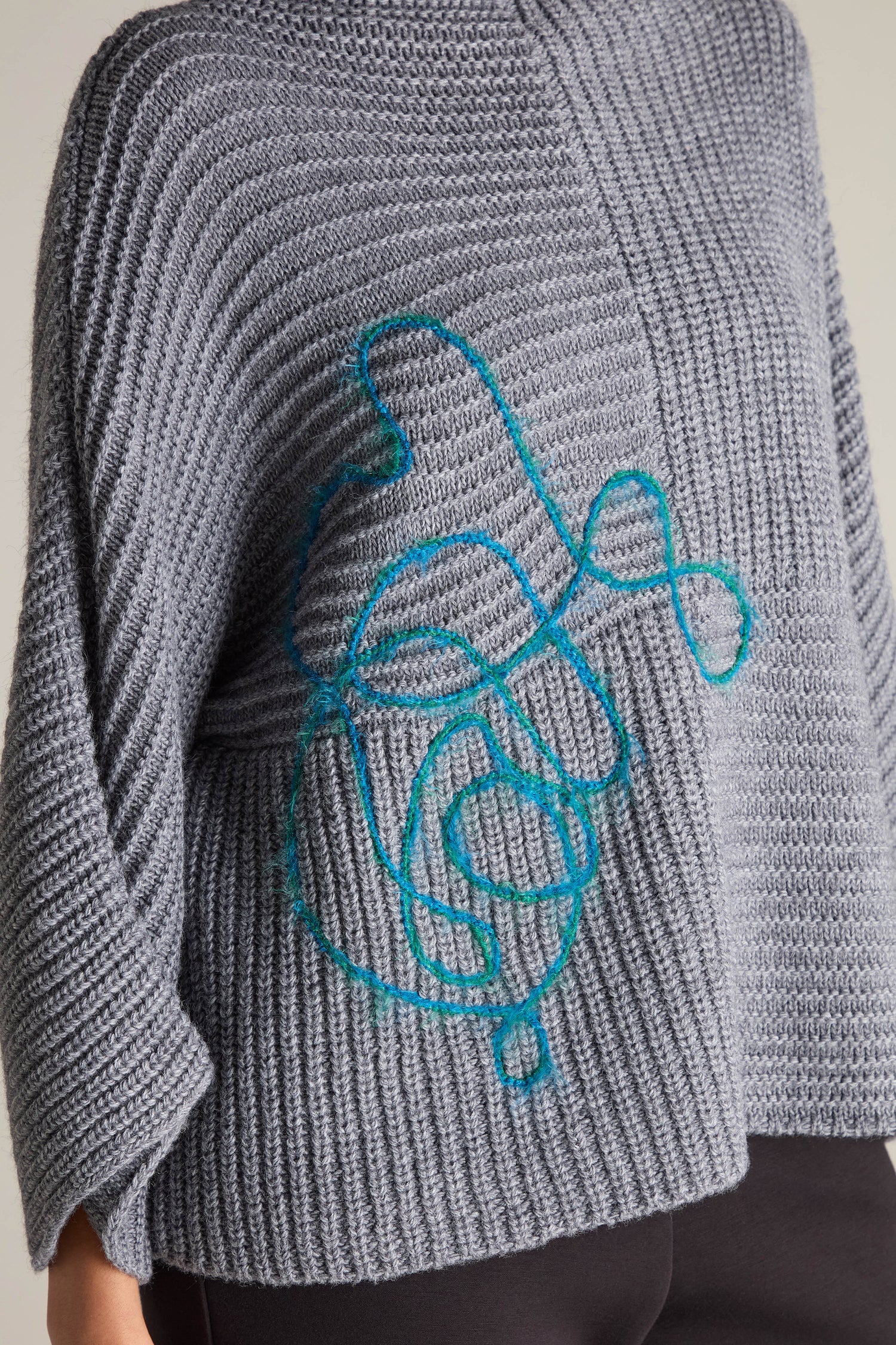 A person wearing the Topstitch Detail Rib Knit, a gray sweater with blue abstract embroidery on the front, made from premium fabric.