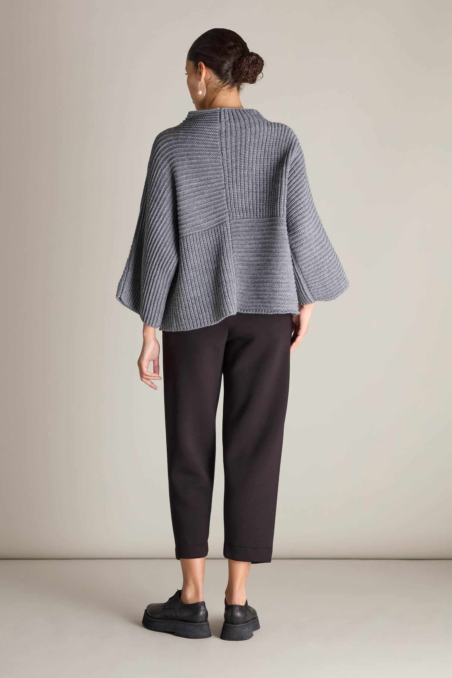 A person is wearing a Topstitch Detail Rib Knit in gray with wide sleeves, crafted from premium fabric, paired with black pants and shoes. They stand against a plain background, facing away from the camera.