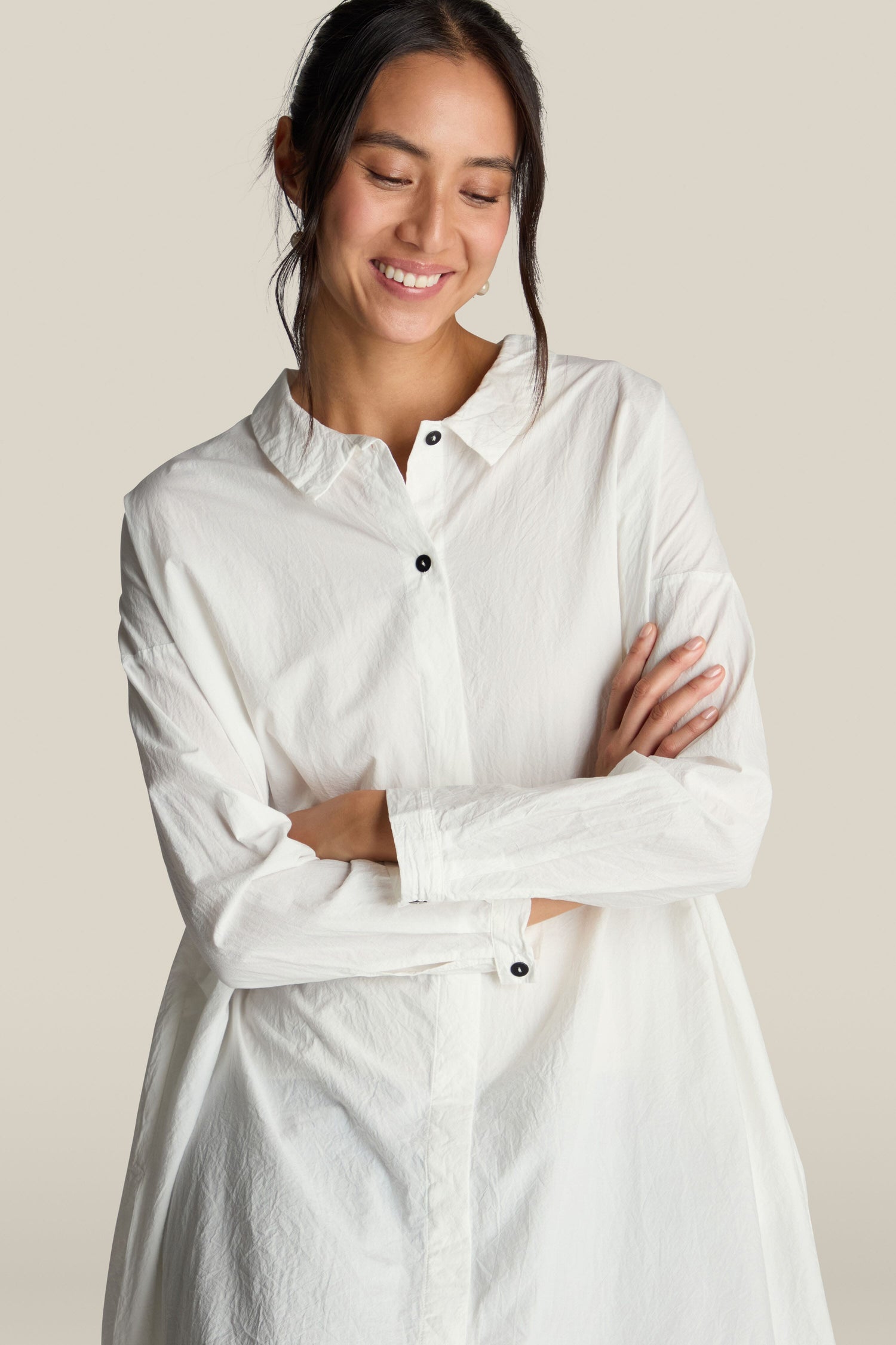 A person wearing the Oversize Shirt in premium cotton white, featuring long sleeves and button-up design, stands with arms crossed, smiling and looking down. The plain, light-colored background enhances an air of relaxed sophistication.