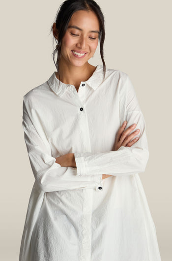 A person wearing the Oversize Shirt in premium cotton white, featuring long sleeves and button-up design, stands with arms crossed, smiling and looking down. The plain, light-colored background enhances an air of relaxed sophistication.