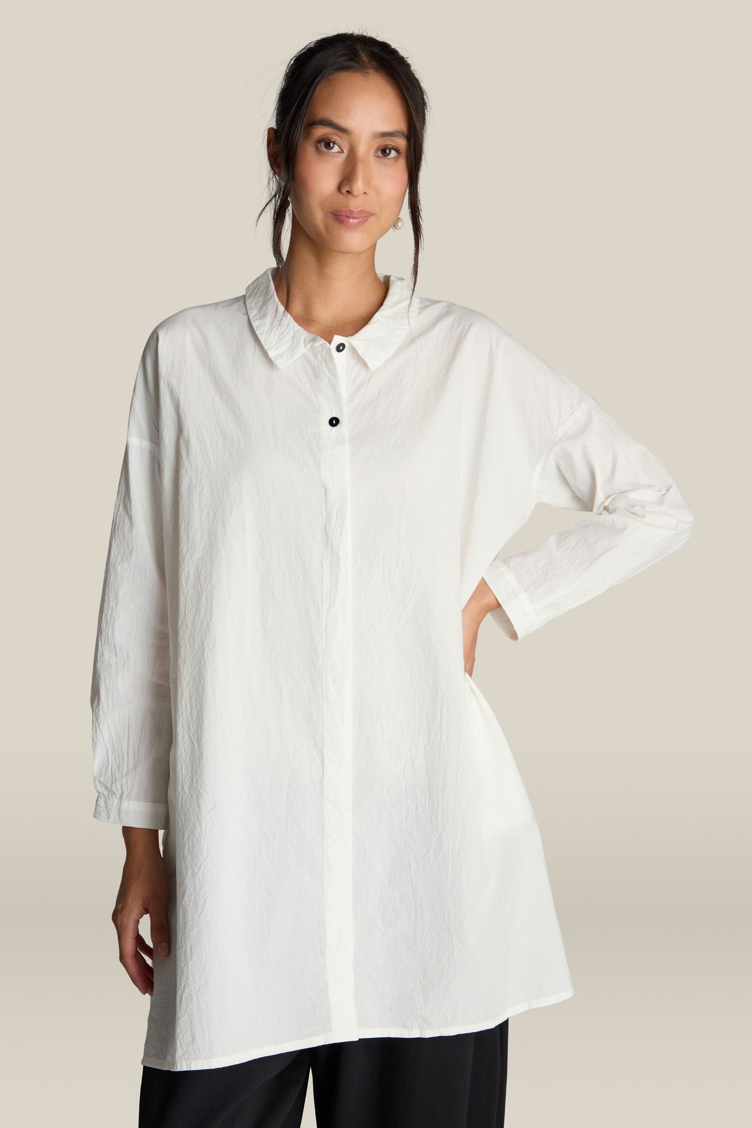 A person is donning the Oversize Shirt, a long-sleeved, oversized white shirt with a collar made from premium cotton. They stand against a plain background, one hand on their hip, exuding an air of relaxed sophistication as they look at the camera.