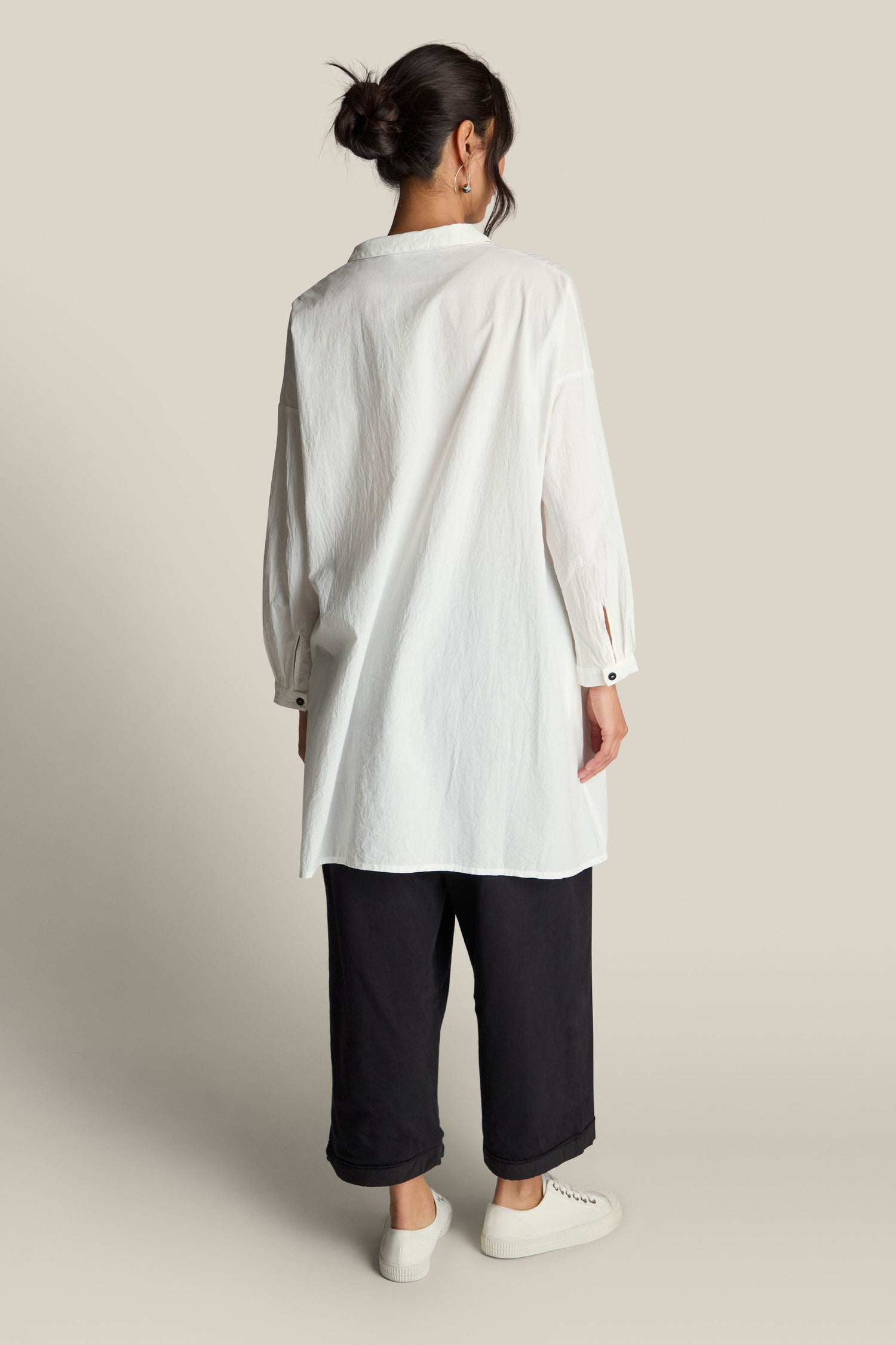 A person stands facing away, wearing an Oversize Shirt in white paired with black loose-fitting pants and white shoes, exuding relaxed sophistication against a plain background.