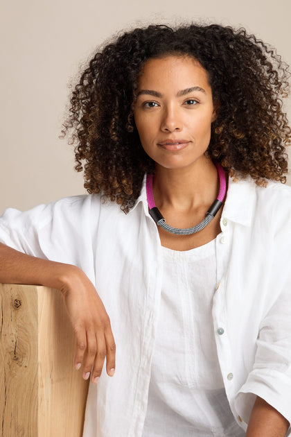 A person with curly hair wearing a white shirt and a modern statement piece—the Short Cord Necklace in pink and black—rests one arm on a wooden surface.