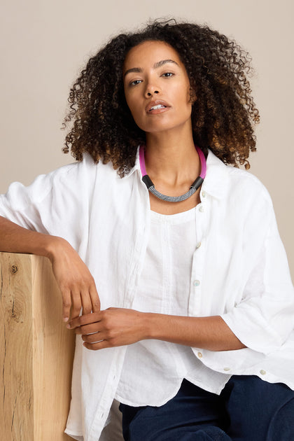 Person with curly hair wearing a white shirt and a trendy Short Cord Necklace, seated against a wooden surface, posing for a photo.