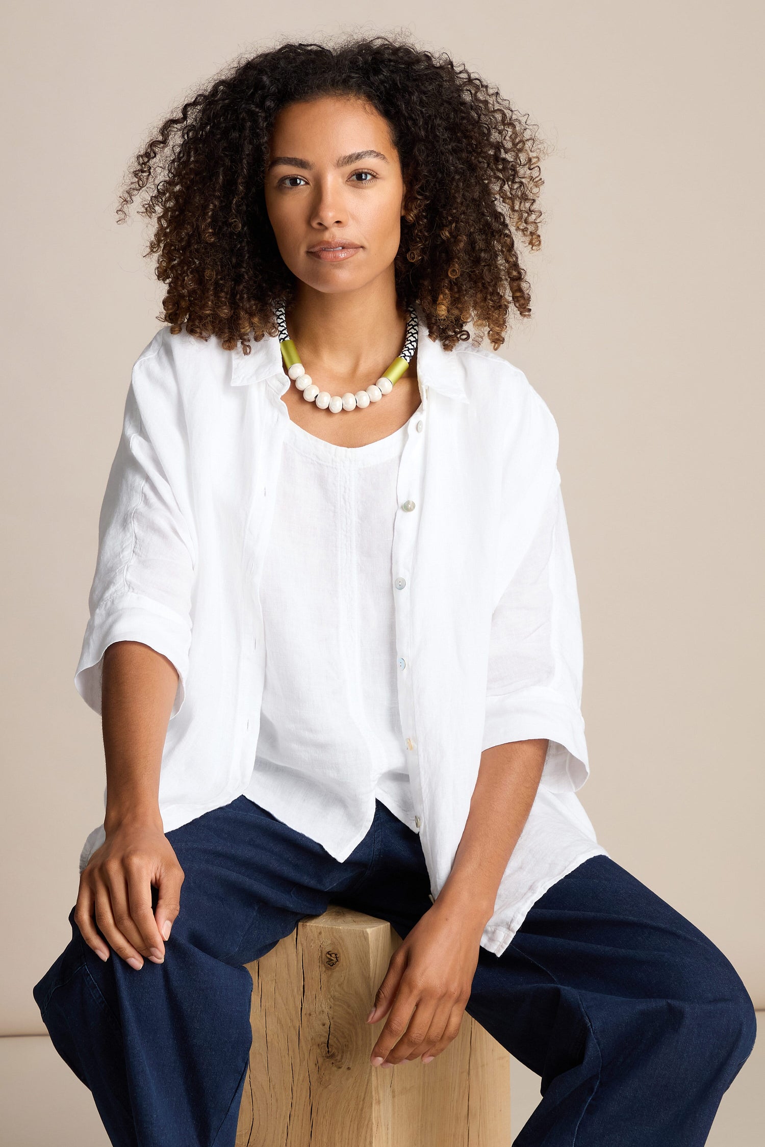 A person with curly hair wears a white shirt, navy pants, and the Milky Way Pearlescent Necklace. Seated on a wooden block against a neutral background, they create a statement look with their contemporary design.