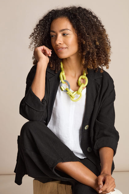 A woman sits on a wooden stool, clad in a black blazer and white shirt. She exudes calm sophistication, looking to the left, her ensemble elevated by the contemporary Zig Zag Hoops Necklace from Christina Brampti.