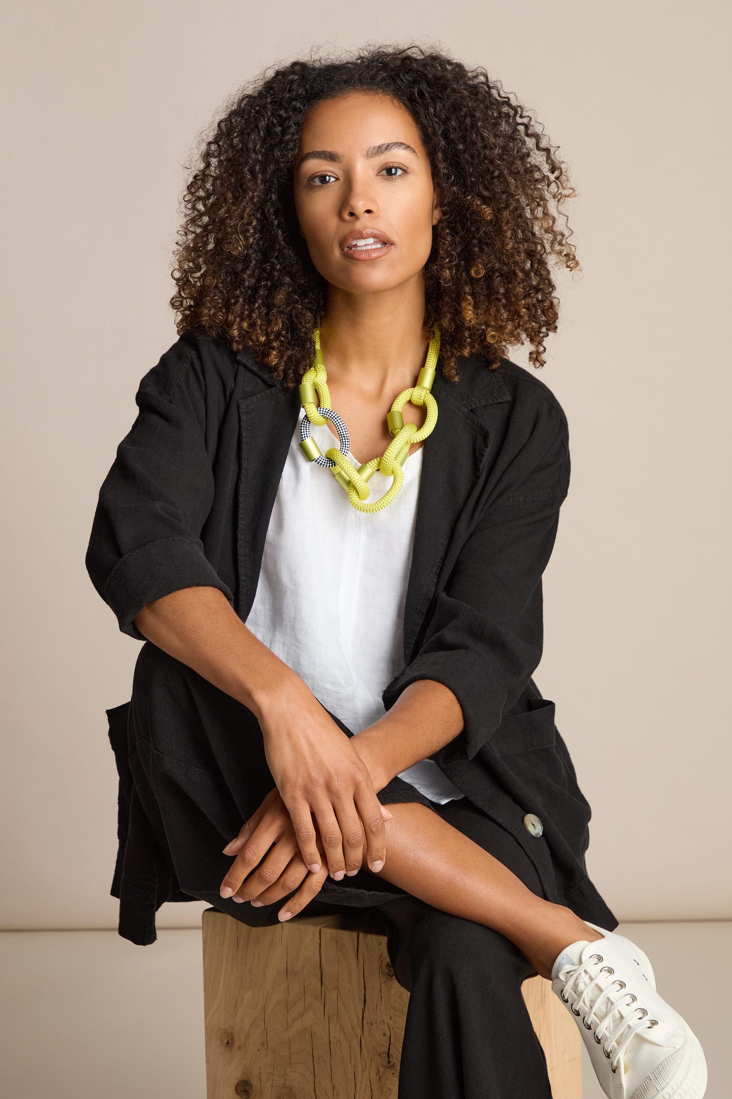 A person with curly hair, wearing a black jacket, white shirt, a yellow Zig Zag Hoops Necklace by Christina Brampti, black pants, and white sneakers, sits on a wooden crate against a beige background.