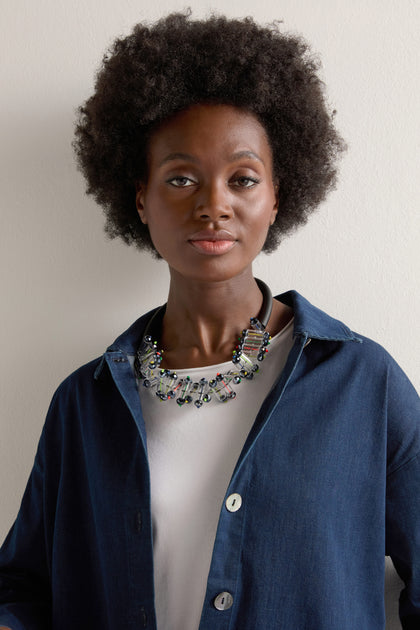 A person with a natural hairstyle is standing against a plain background, wearing a blue shirt over a white top and the Matrix Necklace as a statement accessory.