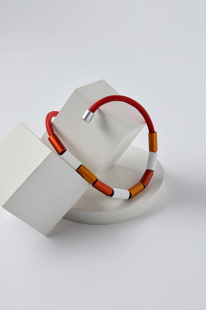 An Aluminium Cylinders Necklace, featuring alternating red, orange, and white rectangular beads, is displayed on a set of white geometric shapes, reminiscent of Christina Brampti's contemporary necklace designs.