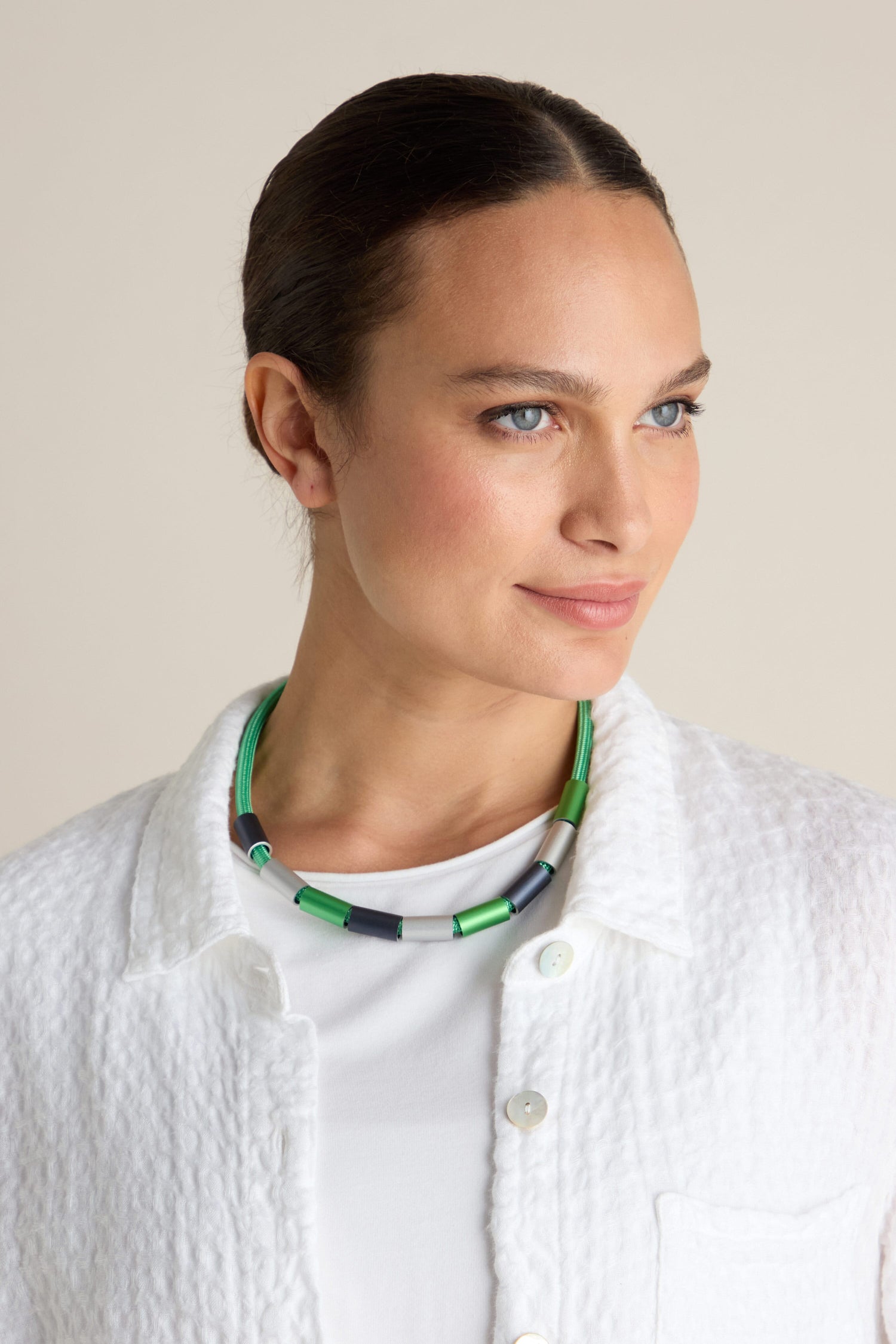 A person wearing a white textured jacket and a white top, accessorized with the Aluminium Cylinders Necklace by Christina Brampti, is looking slightly to the side.