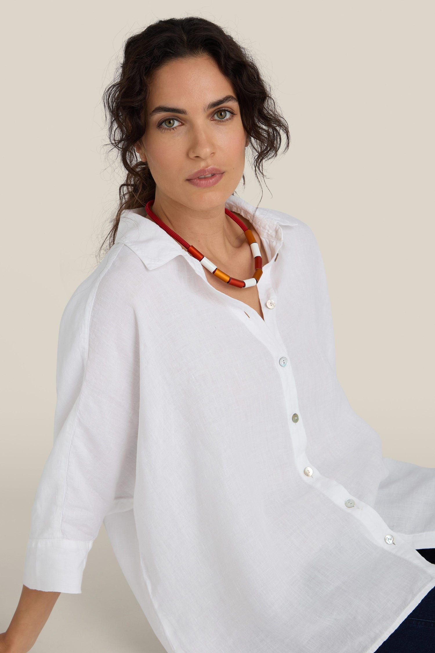 A person with dark curly hair wearing a white button-up shirt and a contemporary necklace, likely the Aluminium Cylinders Necklace, sits and looks at the camera with a neutral expression.