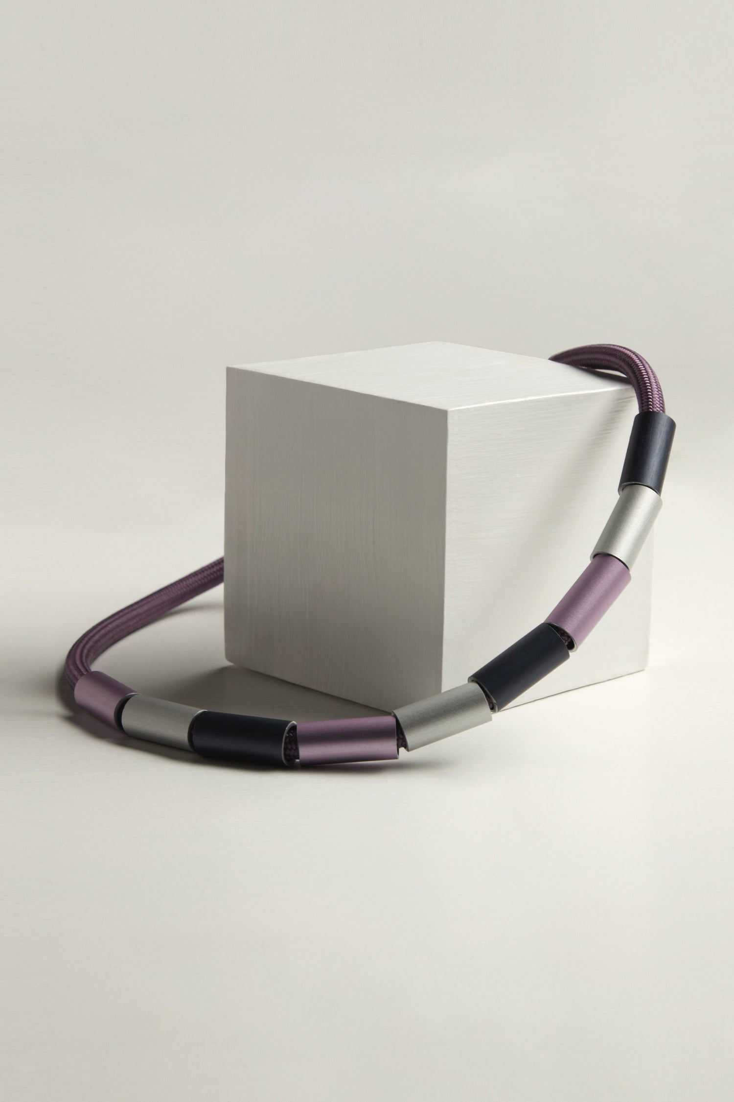 The Aluminium Cylinders Necklace, adorned with purple, silver, and black tubular sections, elegantly drapes over a minimalist white cube, showcasing a sleek and modern statement jewellery design.