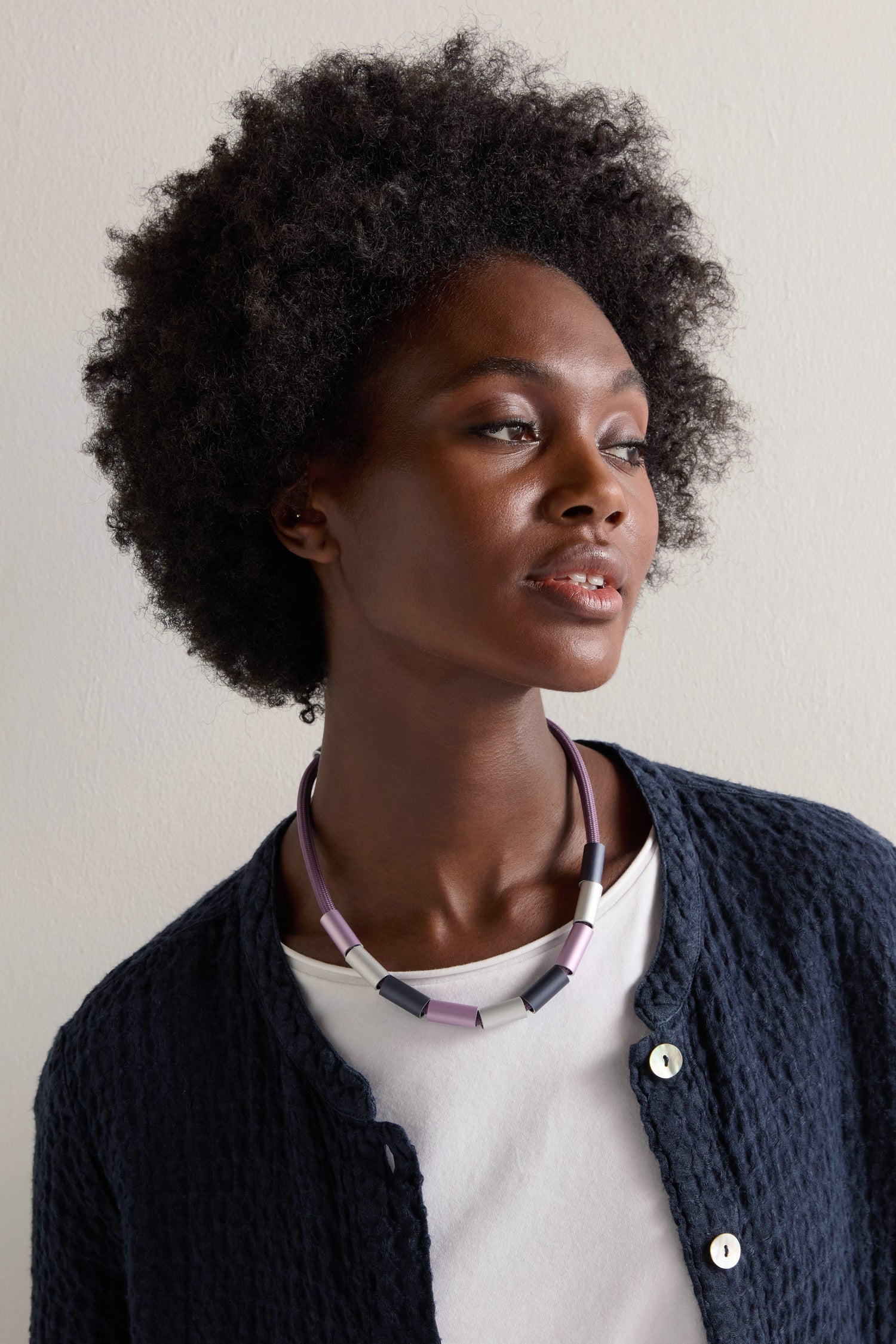 A person with curly hair and a blue textured jacket over a white top wears an Aluminium Cylinders Necklace in purple and black, while looking to the side.