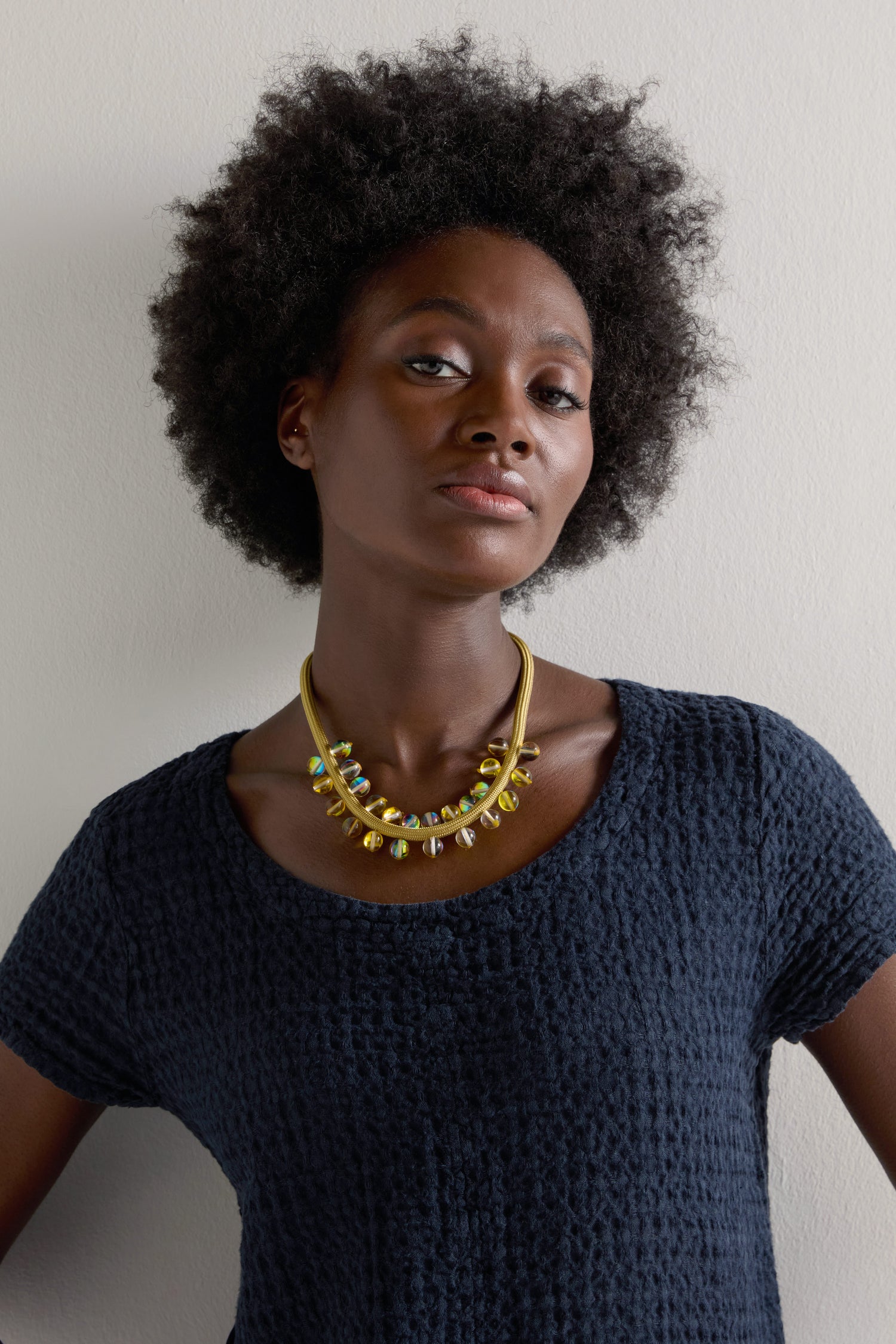 A person with curly hair in a textured navy shirt and an Atmosphere Necklace of iridescent beads stands against a plain background, showcasing contemporary style.