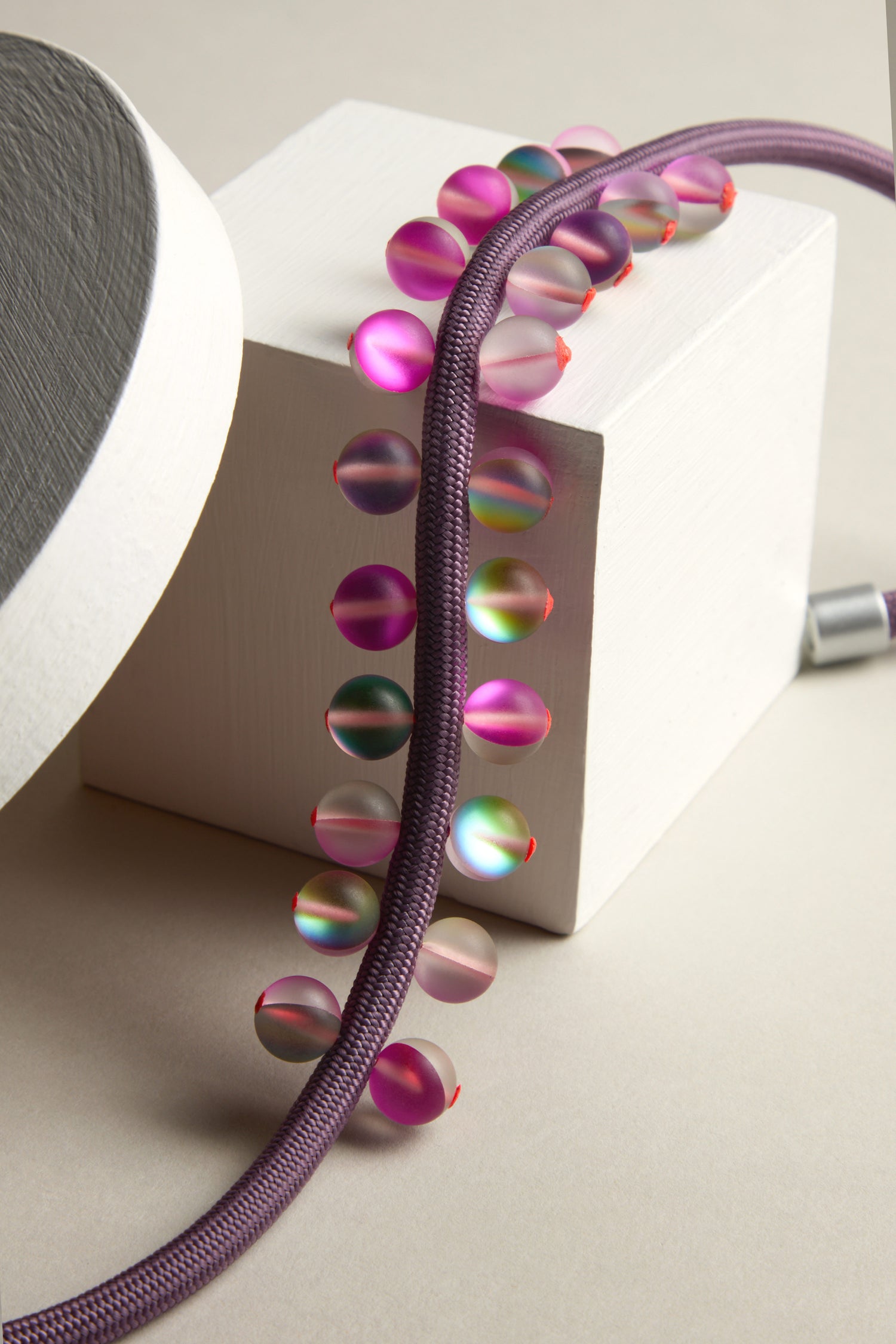 The Atmosphere Necklace combines modern artistry with a geometric arrangement, showcasing iridescent beads on a purple cord elegantly draped over a white cube and curved surface.