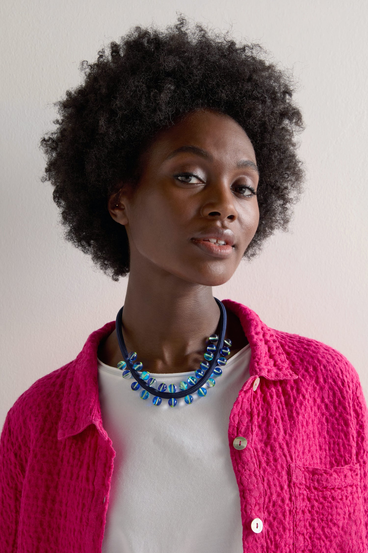 A person with curly hair wears a pink textured shirt, white top, and an Atmosphere Necklace with iridescent beads, standing against a plain background.