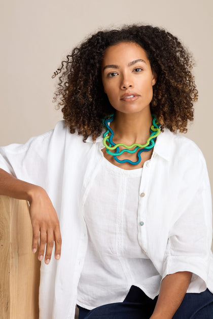 A person with curly hair, adorned in a white shirt and the lively Cord Waves Necklace by Christina Brampti, poses confidently against a beige background, highlighting modern jewelry.