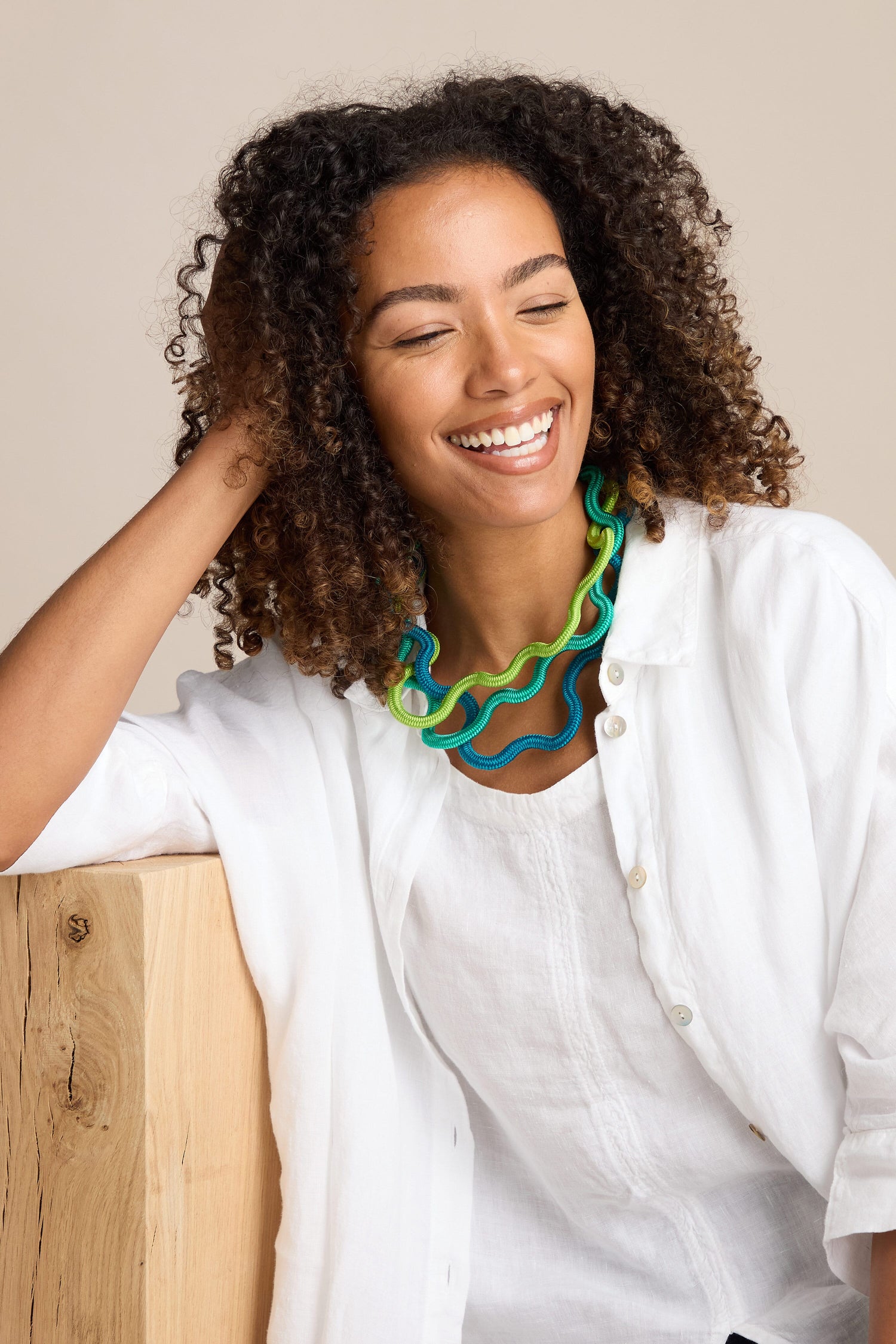 A person with curly hair smiles, wearing a white shirt and a colorful Cord Waves Necklace by Christina Brampti, leaning on a wooden surface.