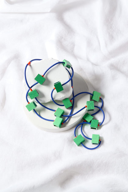 A coiled blue cord adorned with attached green rectangular shapes, reminiscent of the Long Aluminium Cube Necklace by Chrisina Brampti, is artfully arranged on a white circular object against a wrinkled white fabric background. This intriguing setup serves as a bold statement piece.