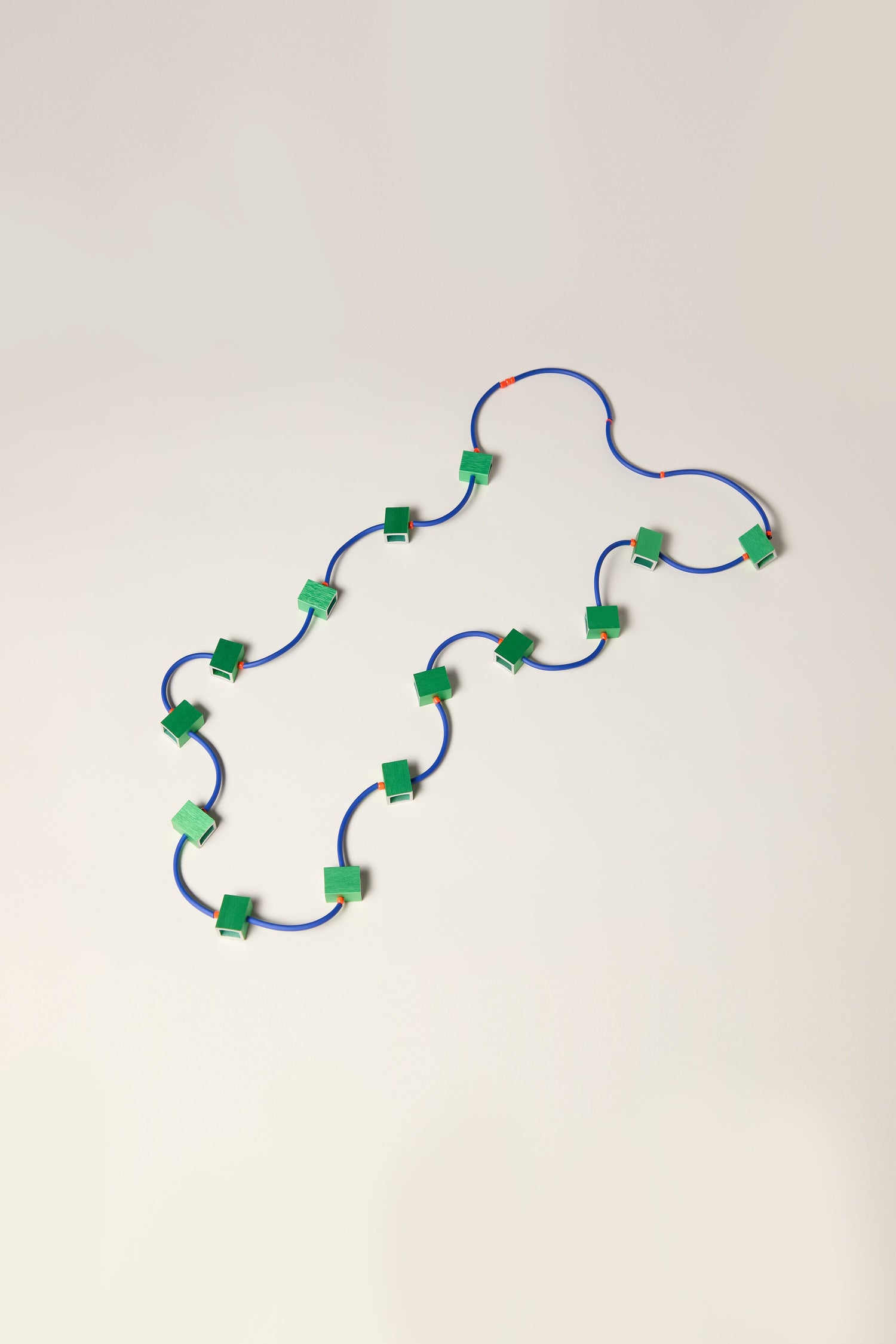 A series of small green rectangles connected by blue wires, arranged in a wave-like pattern on a neutral background, reminiscent of the Long Aluminium Cube Necklace.